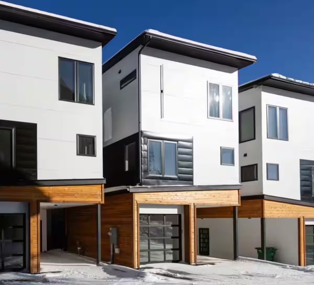 Modern multi-story townhouses with white and black exteriors, wooden accents, large windows, and a snow-covered ground against a clear blue sky.