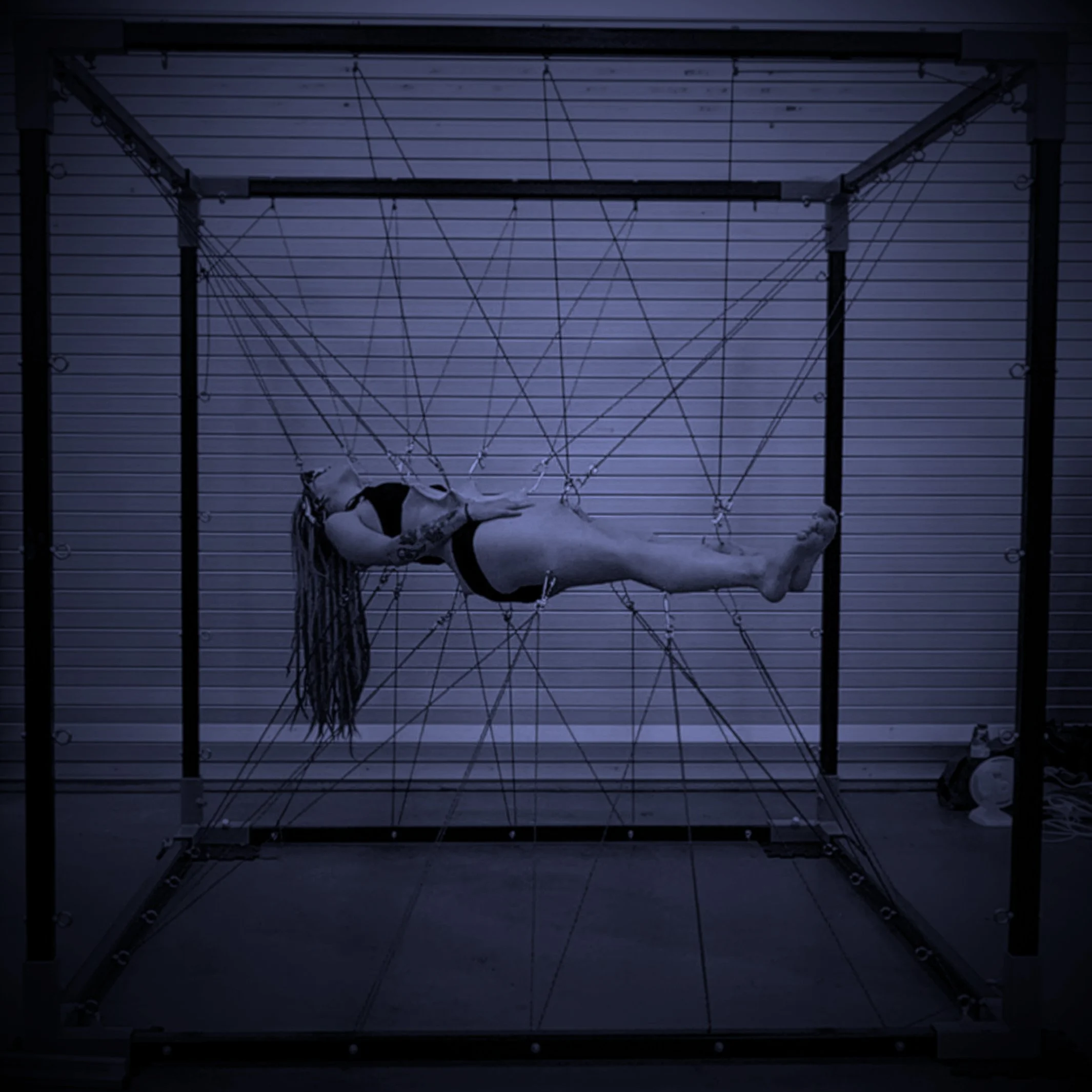 A person with long hair doing a body suspension horizontally in the coma position. inside a geometric metal frame with multiple cords, in a dimly lit room.