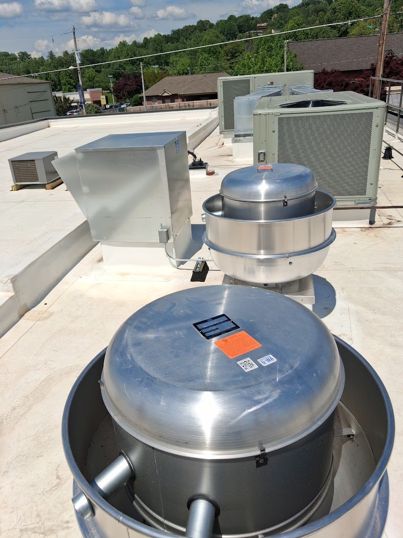 Multiple HVAC units and industrial vents on a building rooftop with a background of trees and houses under a partly cloudy sky.