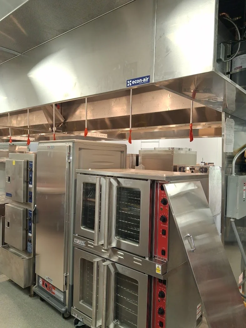 Commercial kitchen with stainless steel ovens, a large exhaust hood, and various kitchen appliances.