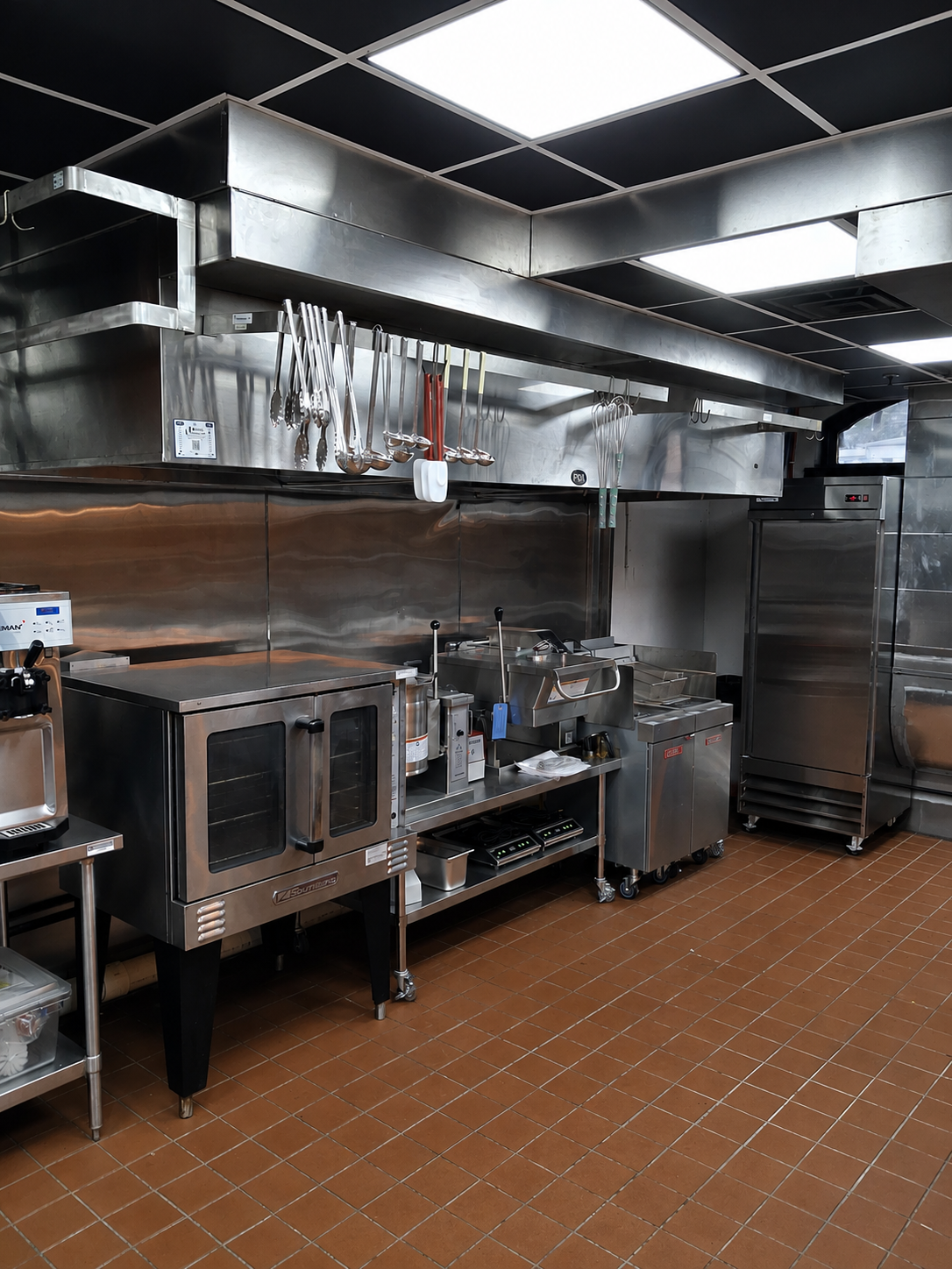 Commercial kitchen with stainless steel appliances, hanging utensils, and brown tile flooring.