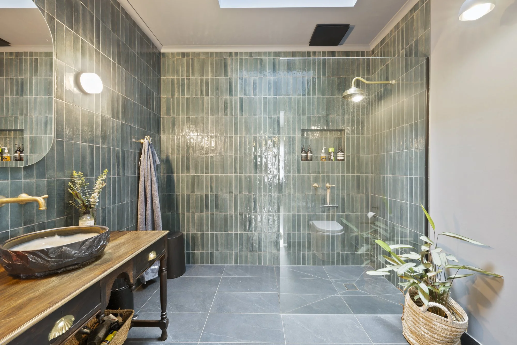 Modern bathroom with green tiled walls, a walk-in shower with a rainfall showerhead, a wooden vanity with a black sink, a mirror, a grey towel hanging on a gold hook, a small plant in a basket, and a gray tile floor.