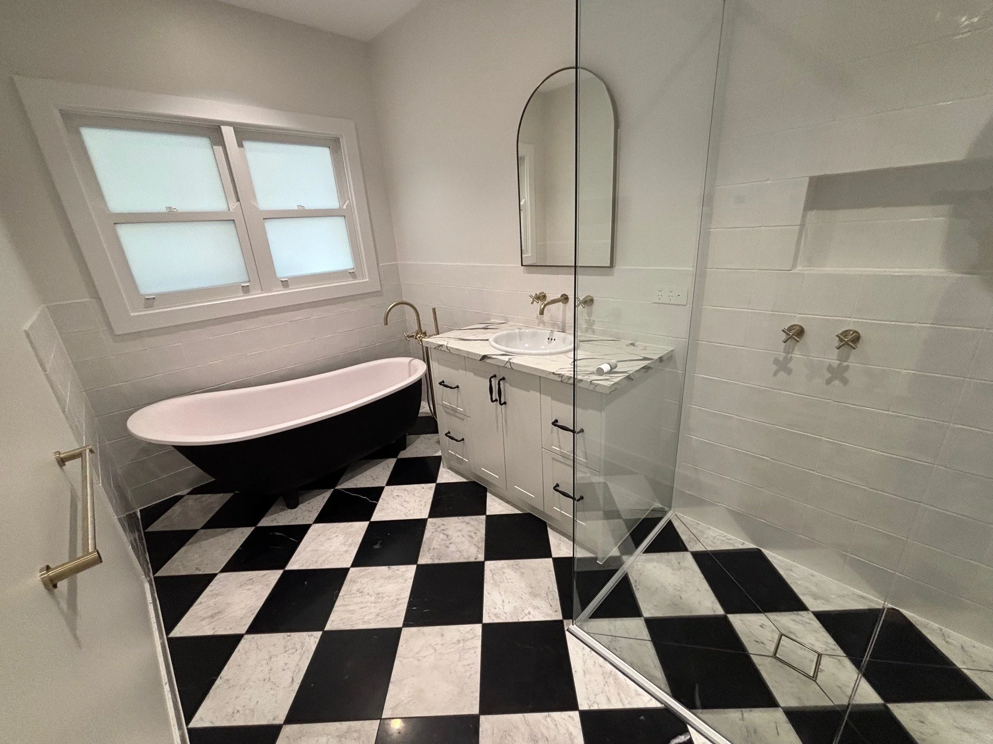 Modern bathroom with black and white checkered marble floor, white vanity with marble countertop, oval mirror, gold fixtures, a bathtub, a window, and a glass shower partition.