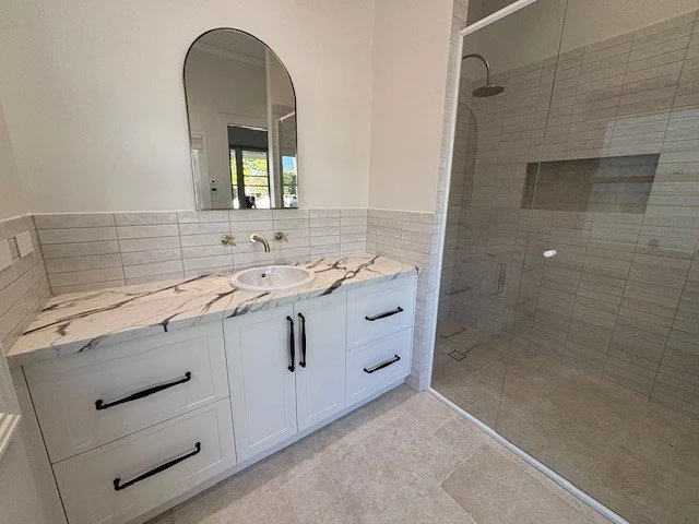 Bathroom with a white vanity with black handles, marble countertop, oval mirror, and a walk-in shower with glass door.