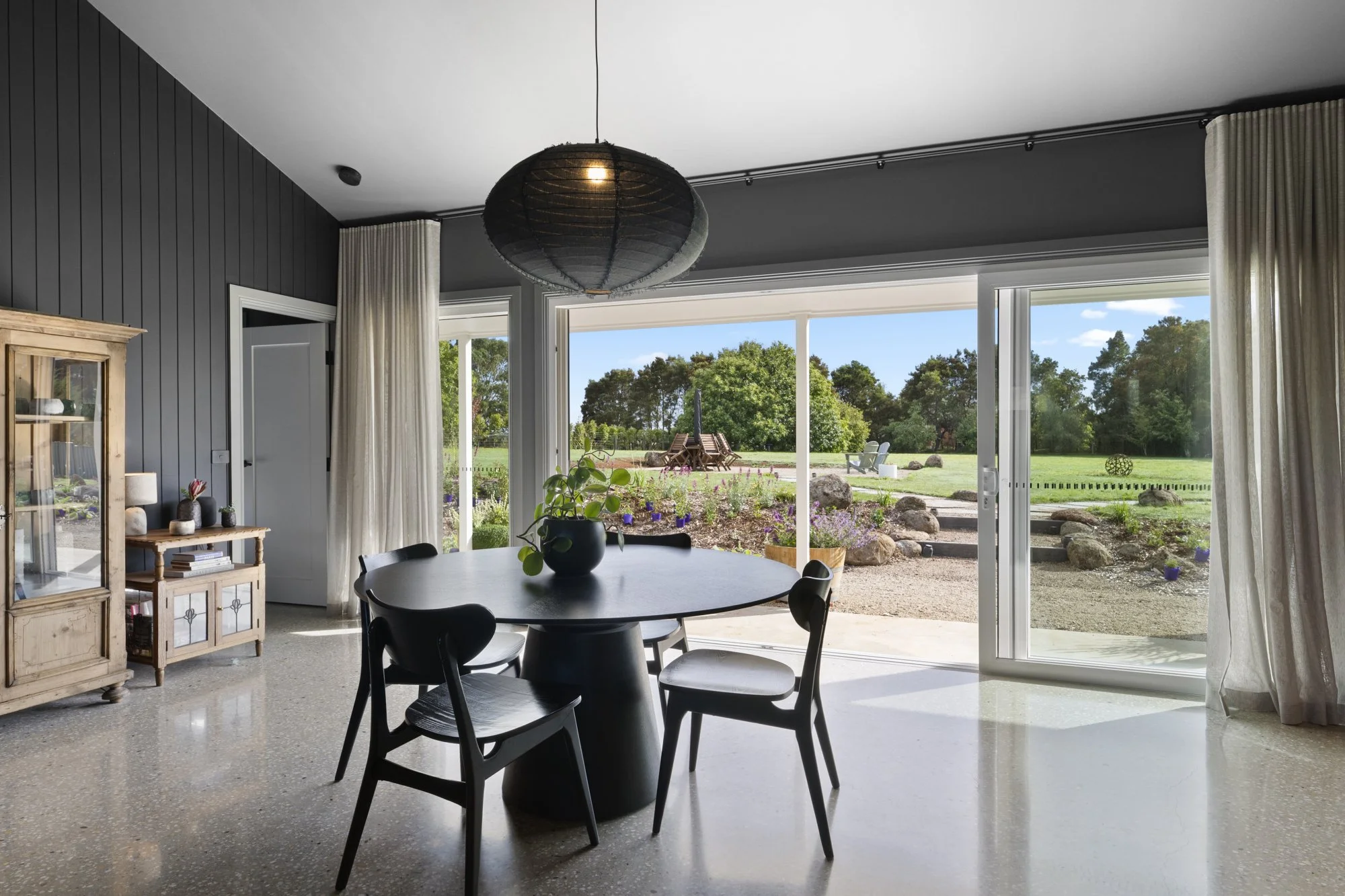 Dining room with black round table, four black chairs, a black pendant light, wooden cabinet, vase with plant, and sliding glass doors leading to garden with rocks, flowers, and trees.