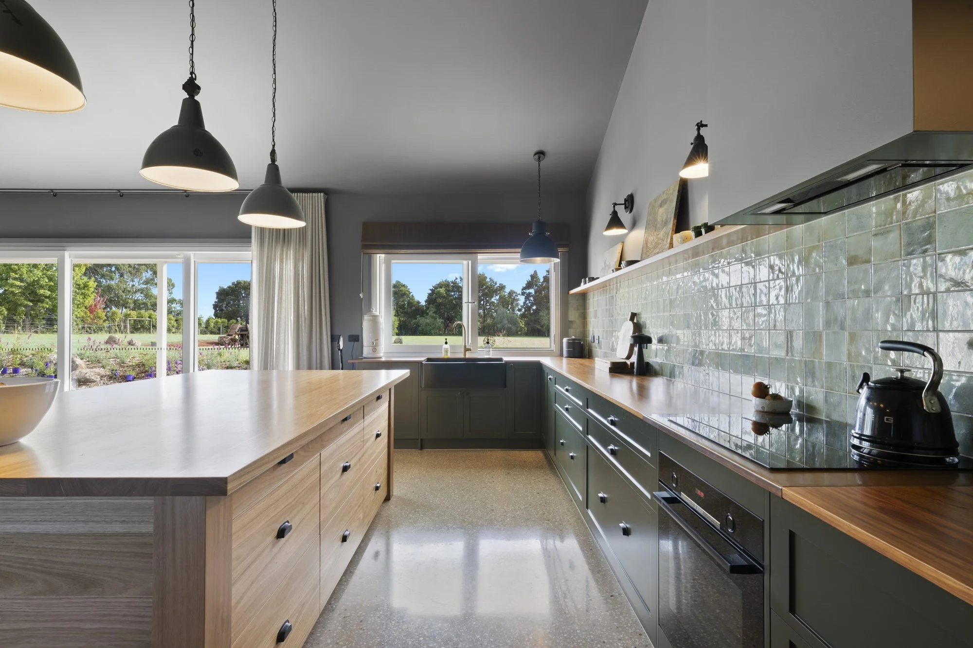 Modern kitchen with green cabinetry, wooden countertops, and a large island. Large windows show a garden outside, and pendant lights hang from the ceiling.