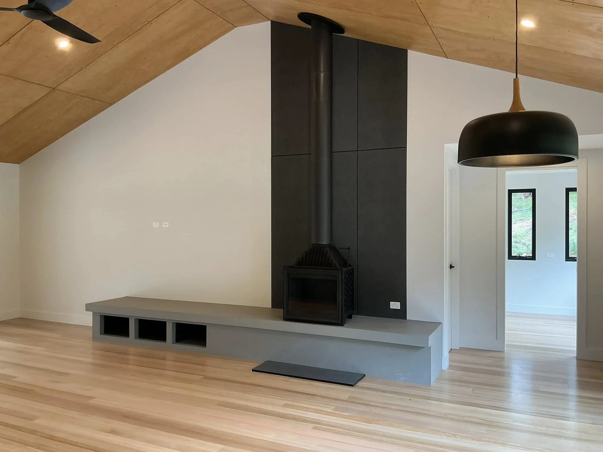 Modern living room with a wood sloped ceiling, a black fireplace with a pipe, and a built-in bench with cubbies. There is a black pendant light and black window frames visible in the background.