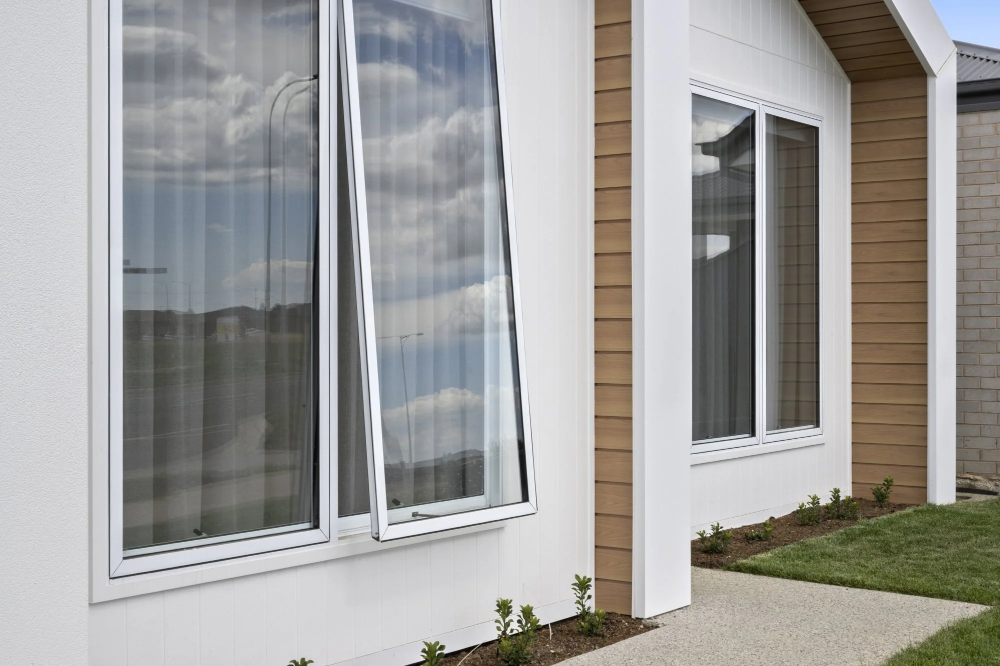 Modern house with large glass windows, some opened, beige and white exterior siding, small landscaped garden with young plants, gravel pathway, and a clear sky with clouds reflected in the windows.