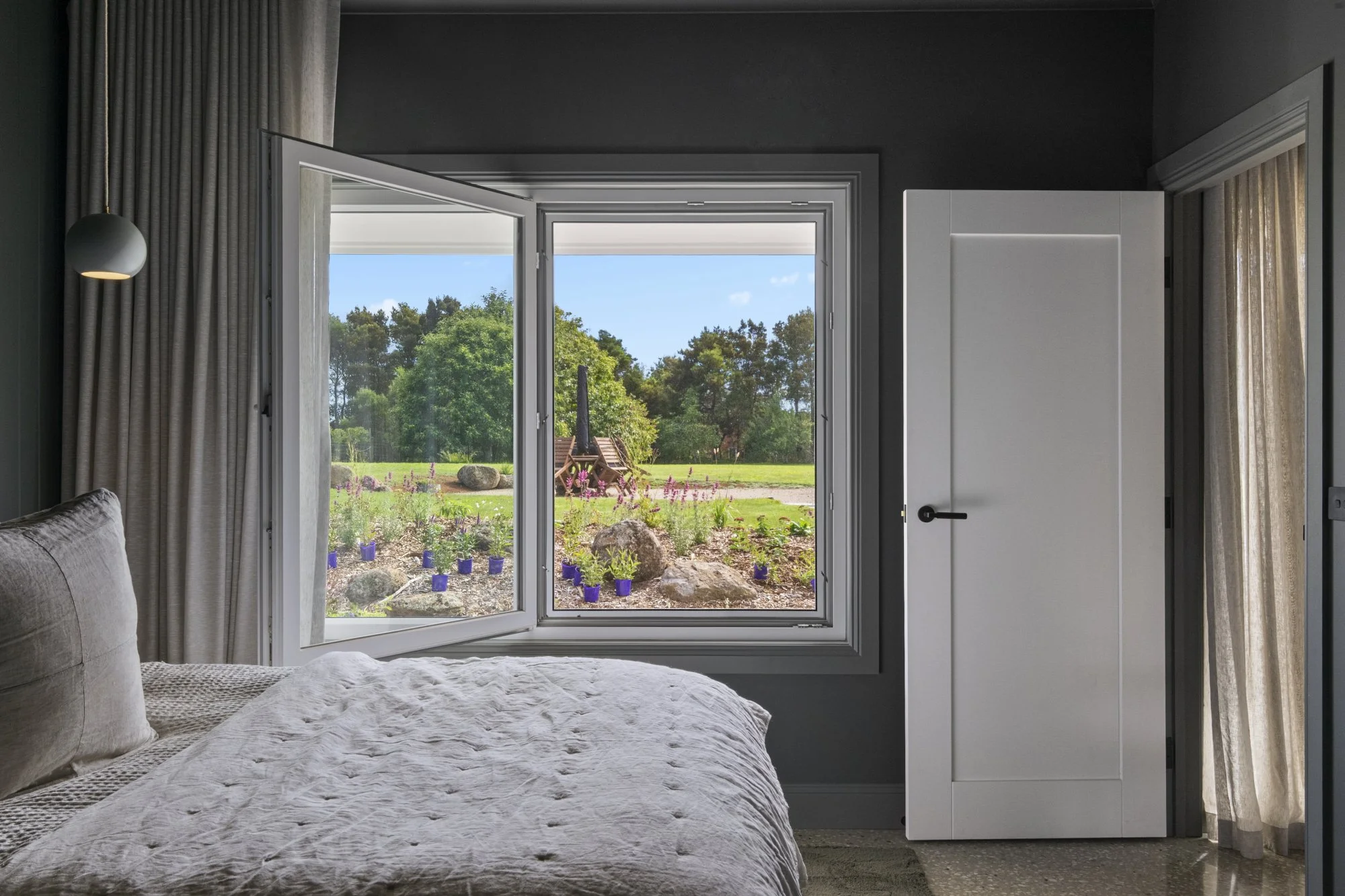 Bedroom with open window overlooking a garden with rocks, plants in purple pots, and trees in the background.