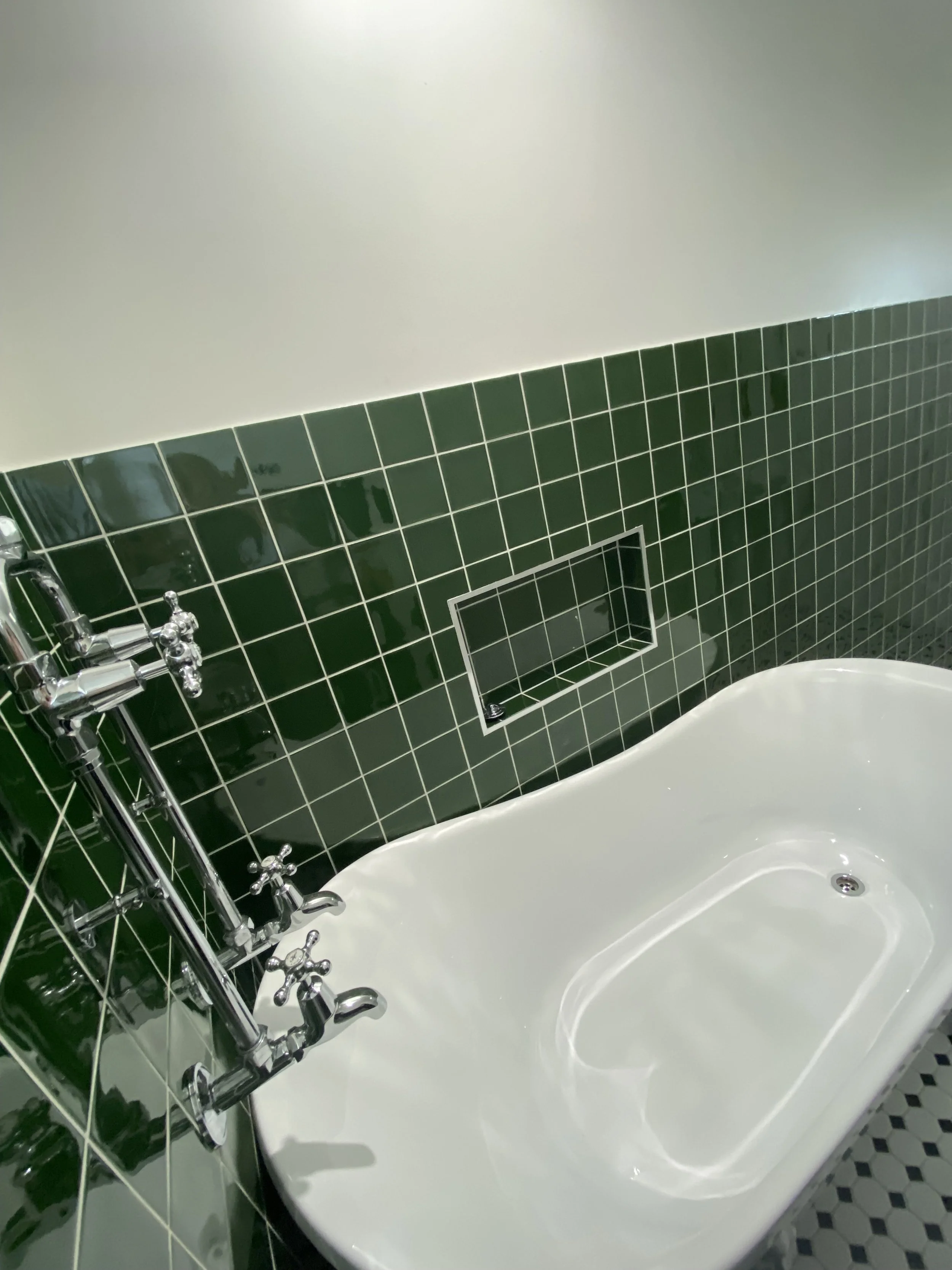 A vintage white bathtub with brass fixtures against a green tiled wall in a bathroom.