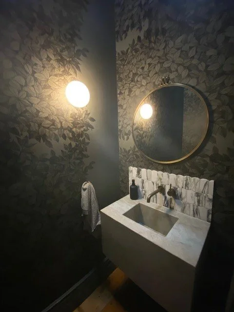 Bathroom with dark leaf-patterned wallpaper, round wall mirror, wall-mounted light fixture, white marble sink, black soap dispenser, and towel hanging on a hook.