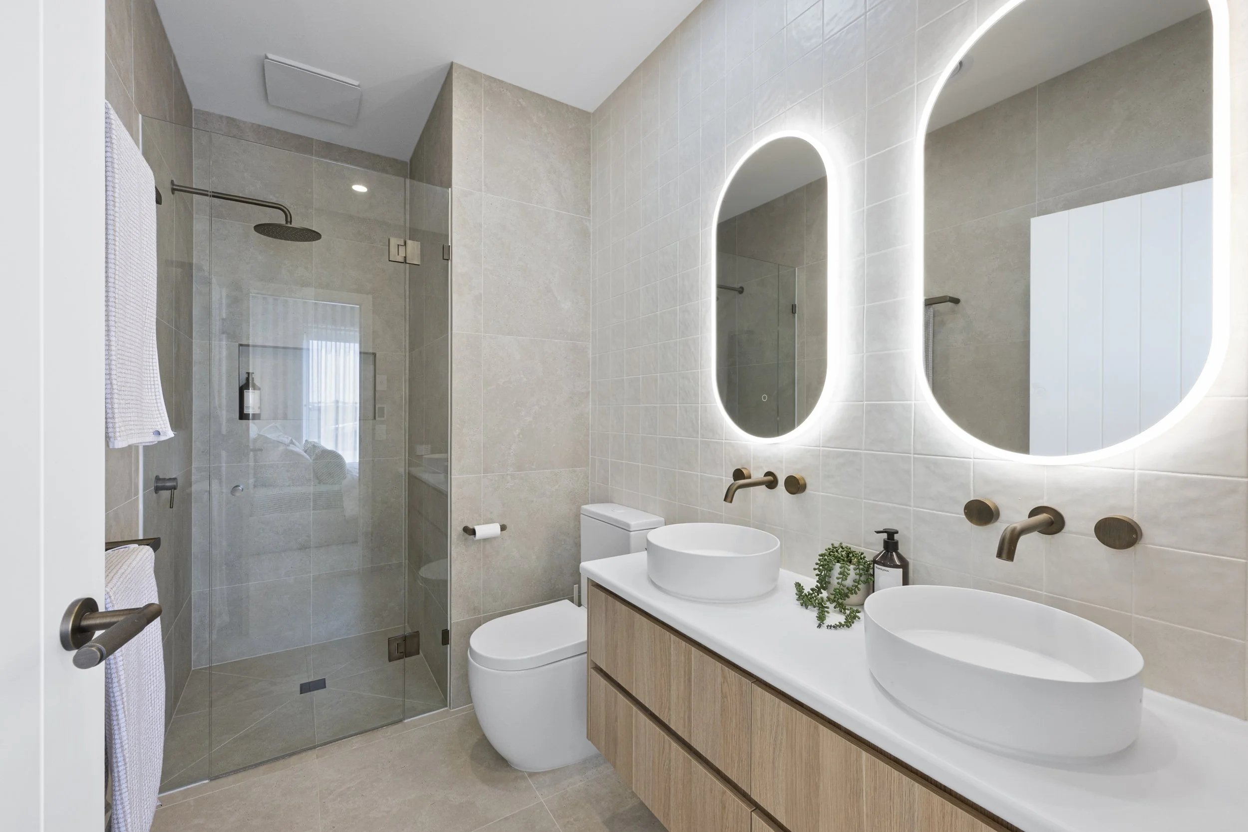 Modern bathroom with double oval sinks, backlit mirrors, beige tiles, glass shower enclosure, and white toilet.