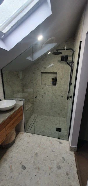 A modern bathroom shower with beige marble walls and floor, a built-in niche, a black showerhead and controls, and a partial view of a wooden vanity with a white vessel sink.