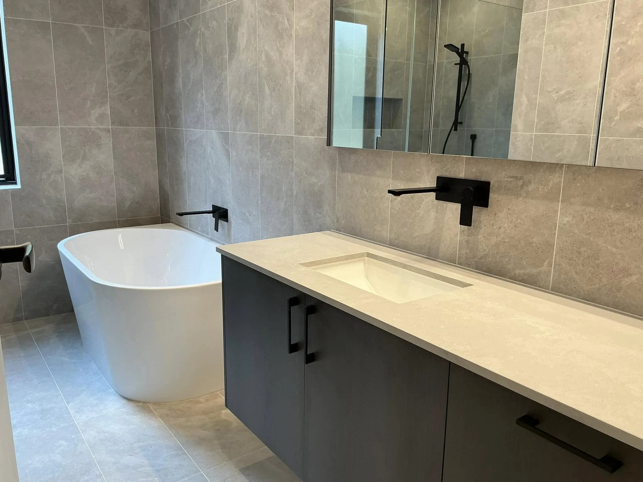 Modern bathroom with gray tiled walls and flooring, a white bathtub, a gray vanity with black handles, a beige countertop with an integrated sink, and black wall-mounted fixtures. A large mirror partially visible above the sink reflects a shower area
