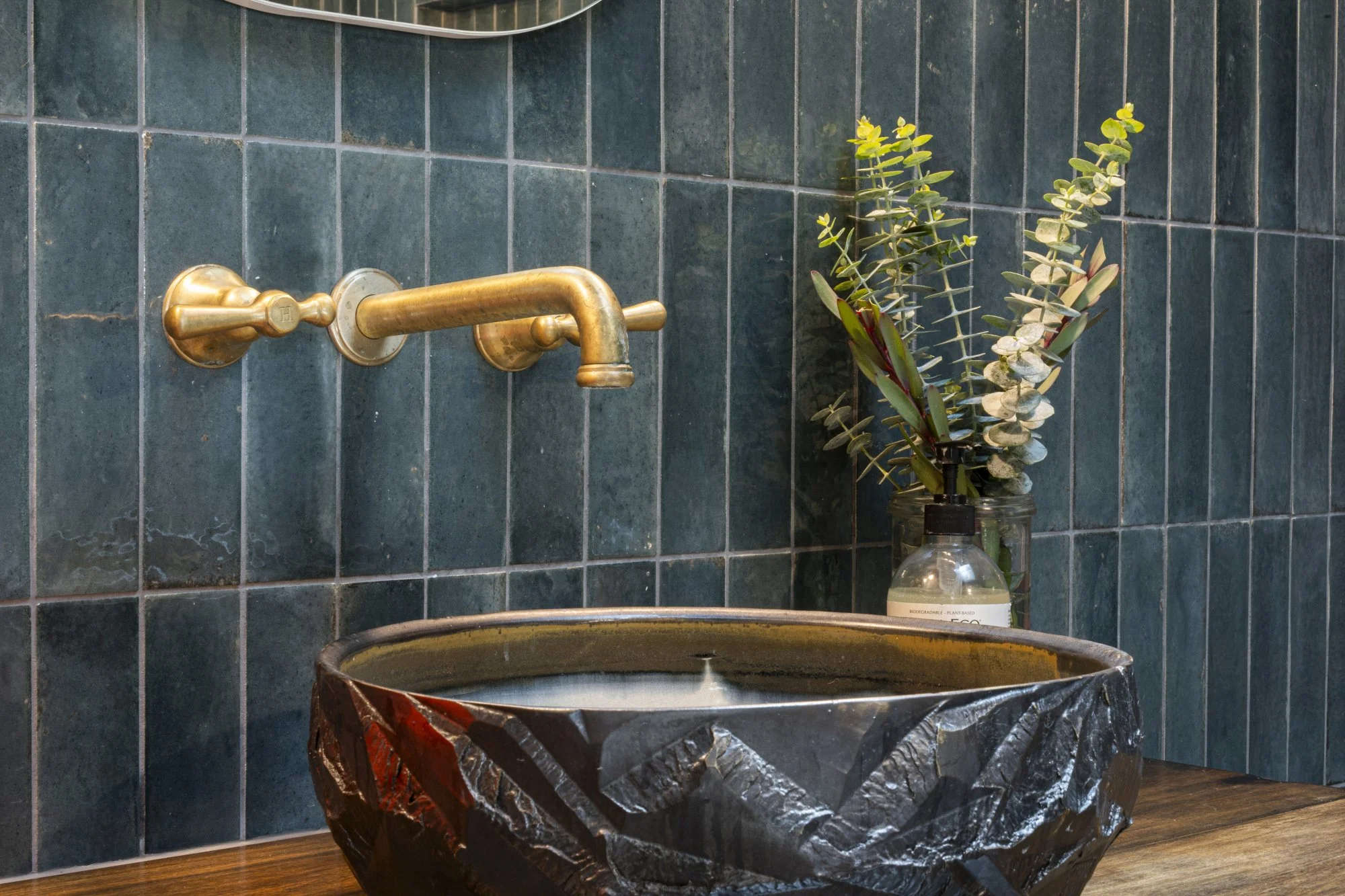 Interior view of a bathroom with dark green tiles, a gold faucet, a decorative black stone sink, and a glass jar with greenery on a wooden countertop.