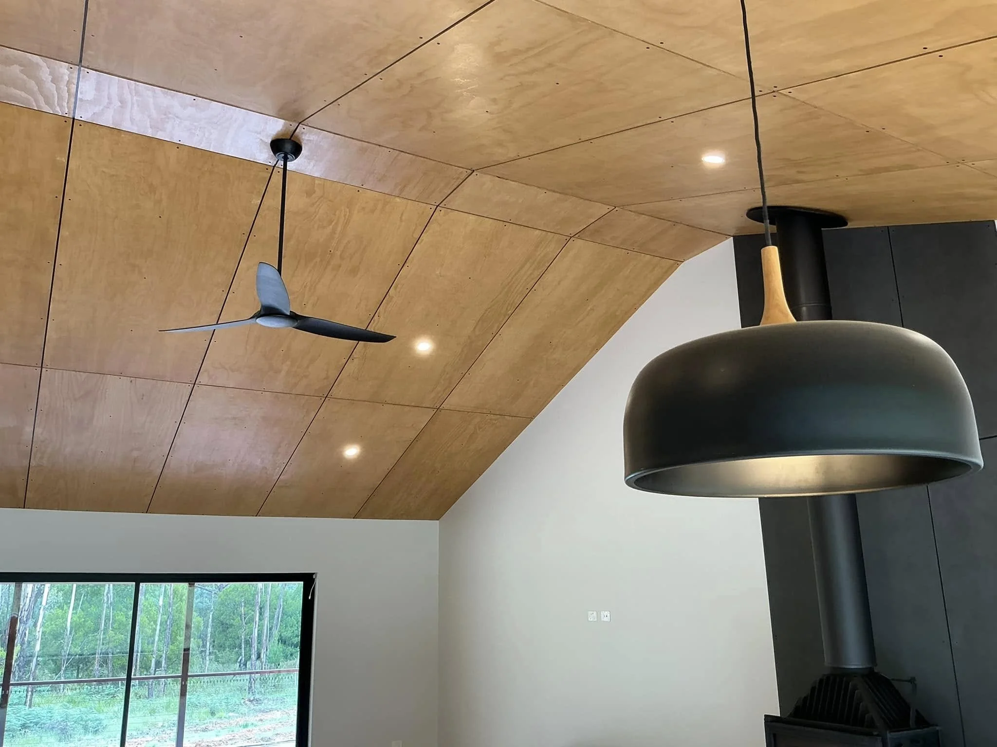 Interior of a room with a wooden ceiling, a black ceiling fan, recessed lighting, large window with green trees outside, and a black stove with a chimney.
