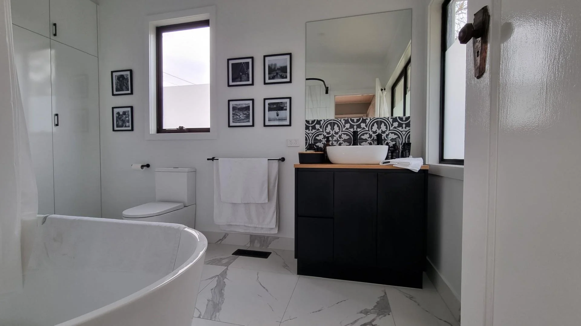 A modern bathroom with black and white decor, a bathtub, a toilet, a sink with black hardware, a large mirror, wall art, and windows for natural light, with marble flooring.