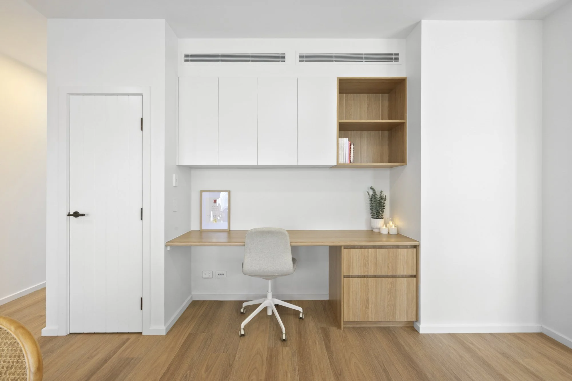 A minimalist home office with a wooden desk, white desk chair, built-in white cabinets, and a small open wooden shelf with a few books. The desk has a framed picture and potted plants with candles. The room has light wooden flooring and white walls.
