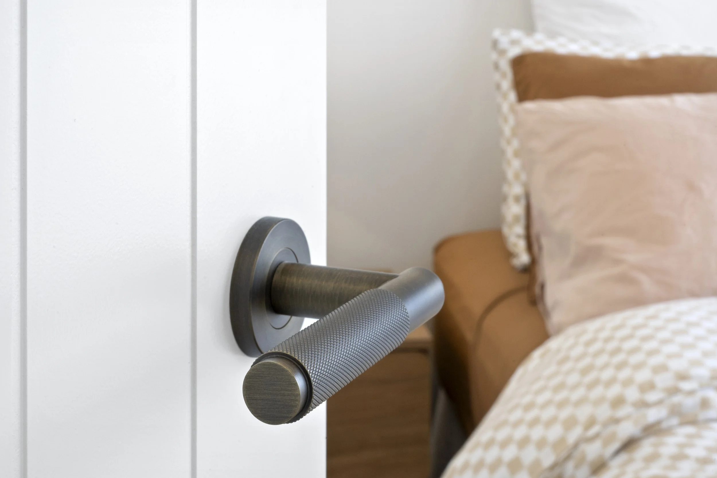 Close-up of a modern metallic door handle on a white door, with part of a bed and pillows visible in the background.