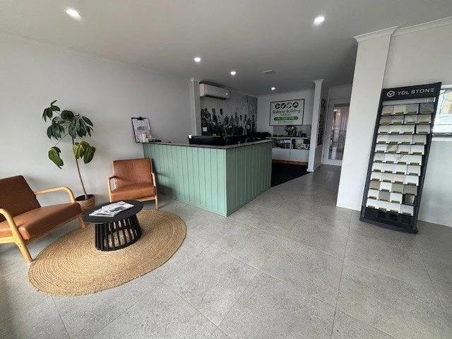 Lobby area with two brown armchairs, a round coffee table on a natural fiber rug, a potted plant, and a reception desk with a light green panel. In the background, there is a display rack, a sign, and a hallway leading to other rooms.