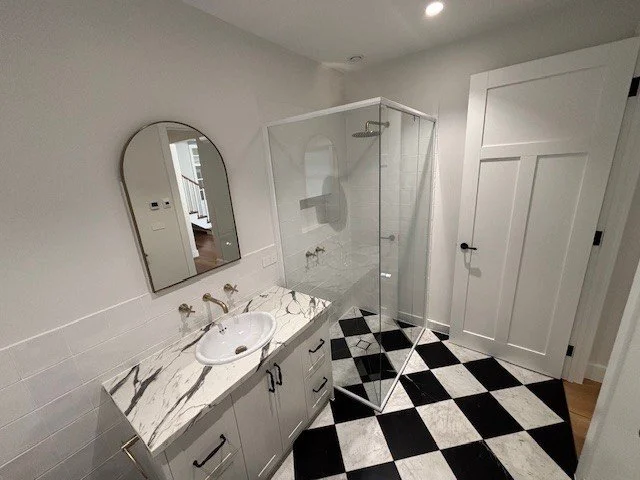 Bathroom with a marble countertop vanity, oval mirror, shower with glass door, and black-and-white checkered floor tiles.