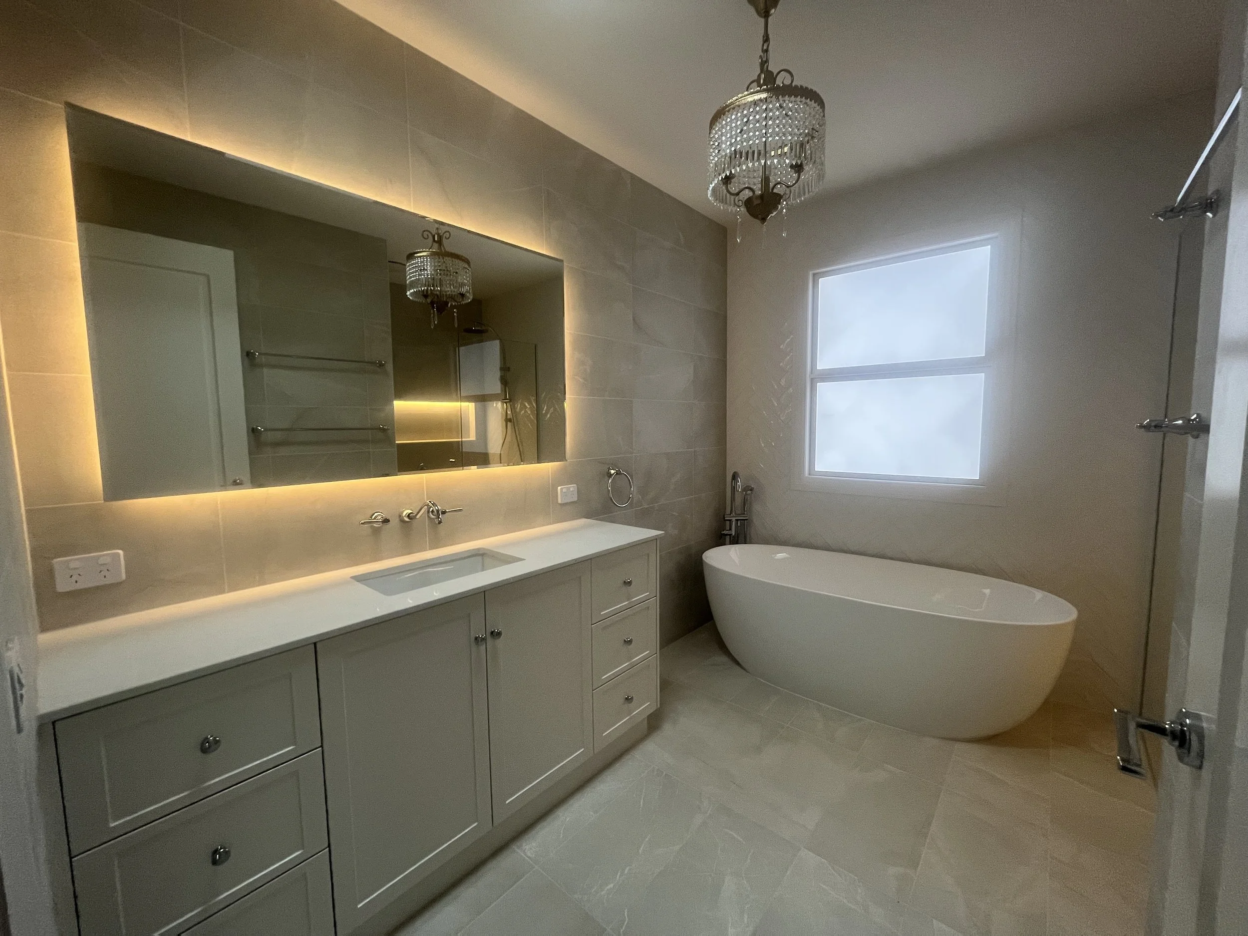 Modern bathroom with a large backlit mirror, white vanity with sink, a freestanding bathtub next to a window, and a chandelier hanging from the ceiling.