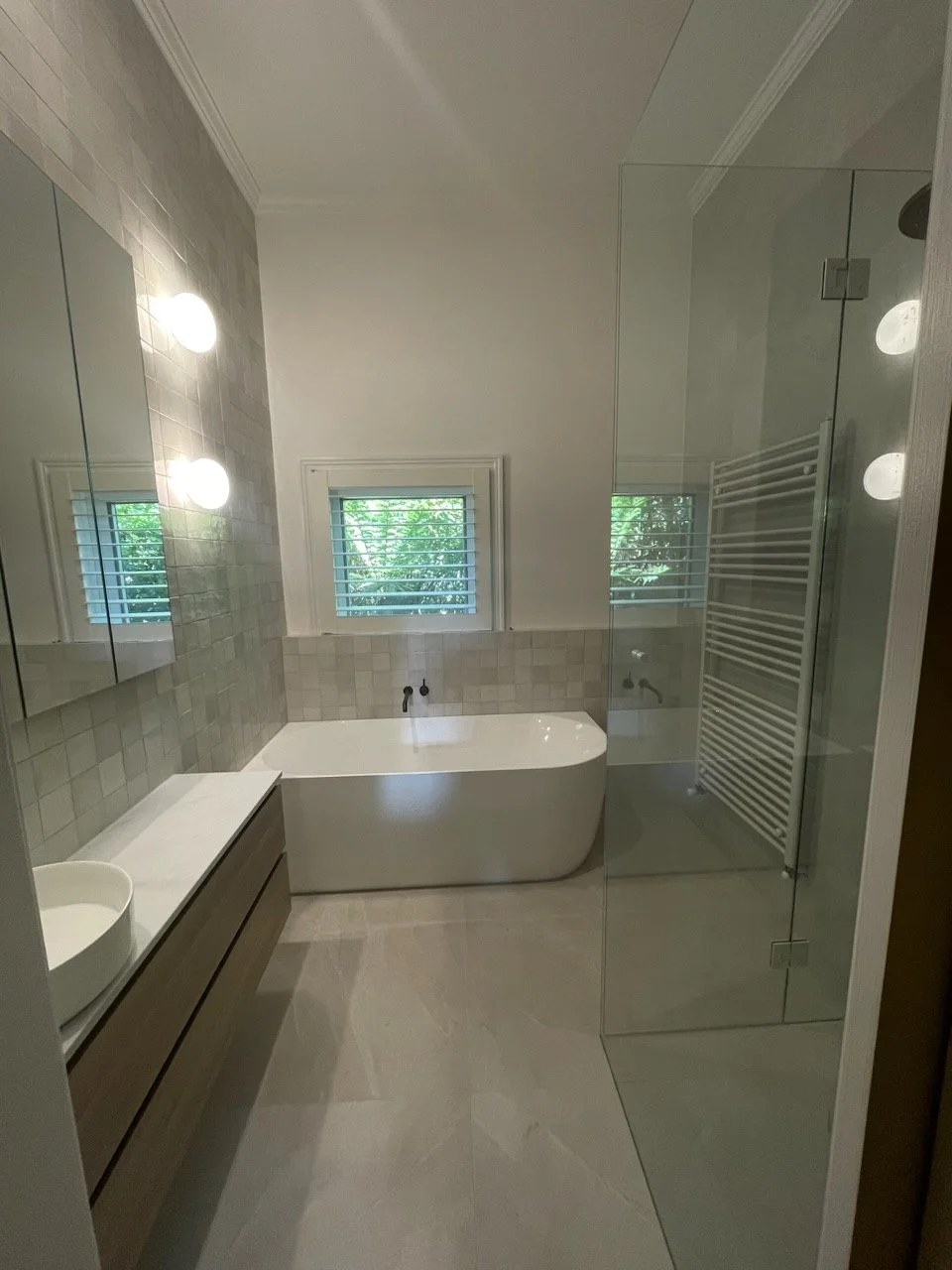 Modern bathroom with a bathtub, a glass-enclosed shower, a small window with blinds, and a white countertop with a vessel sink.