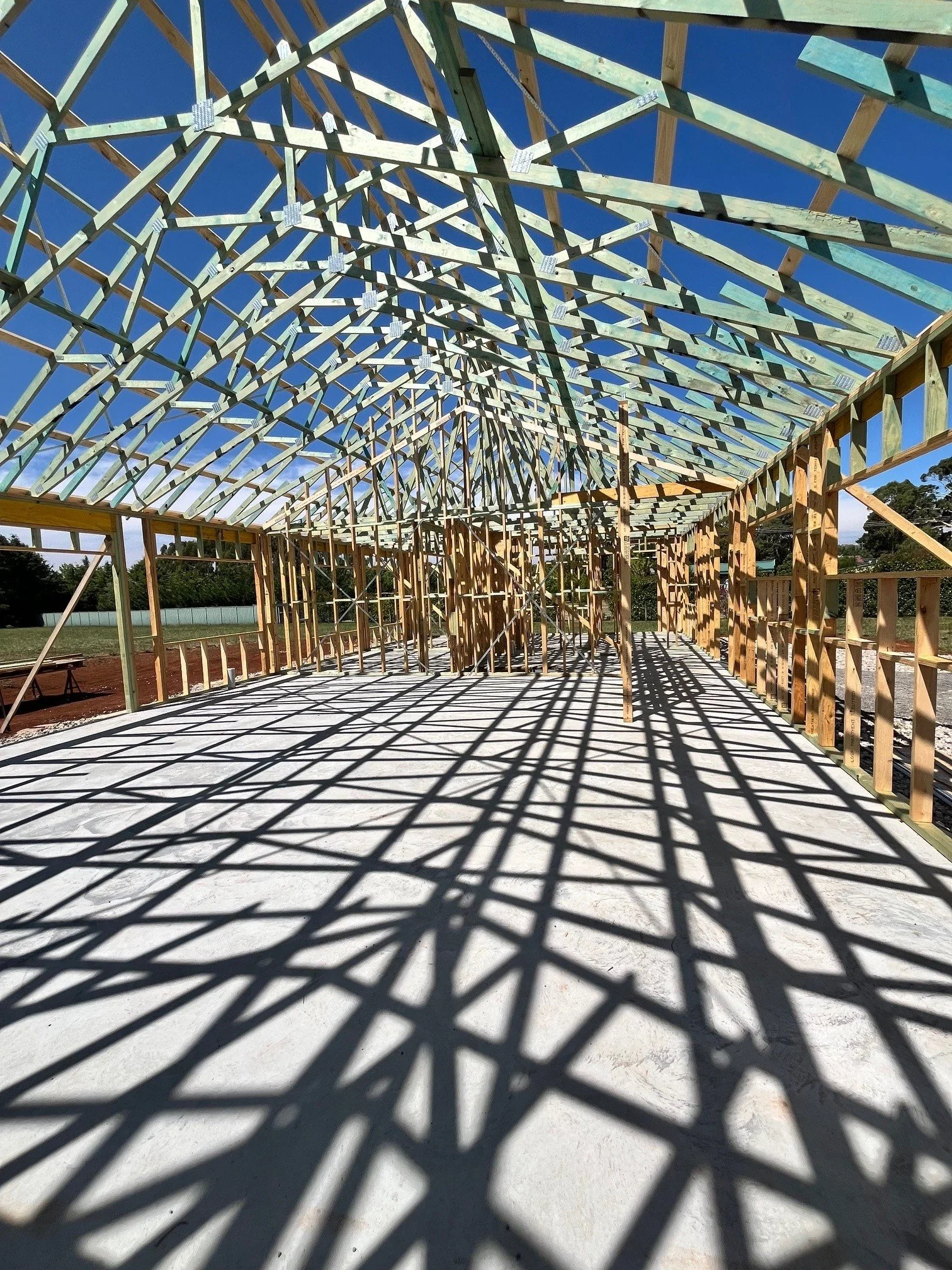 Construction site with wooden framing for a building, casting shadows on the concrete floor under a bright blue sky.