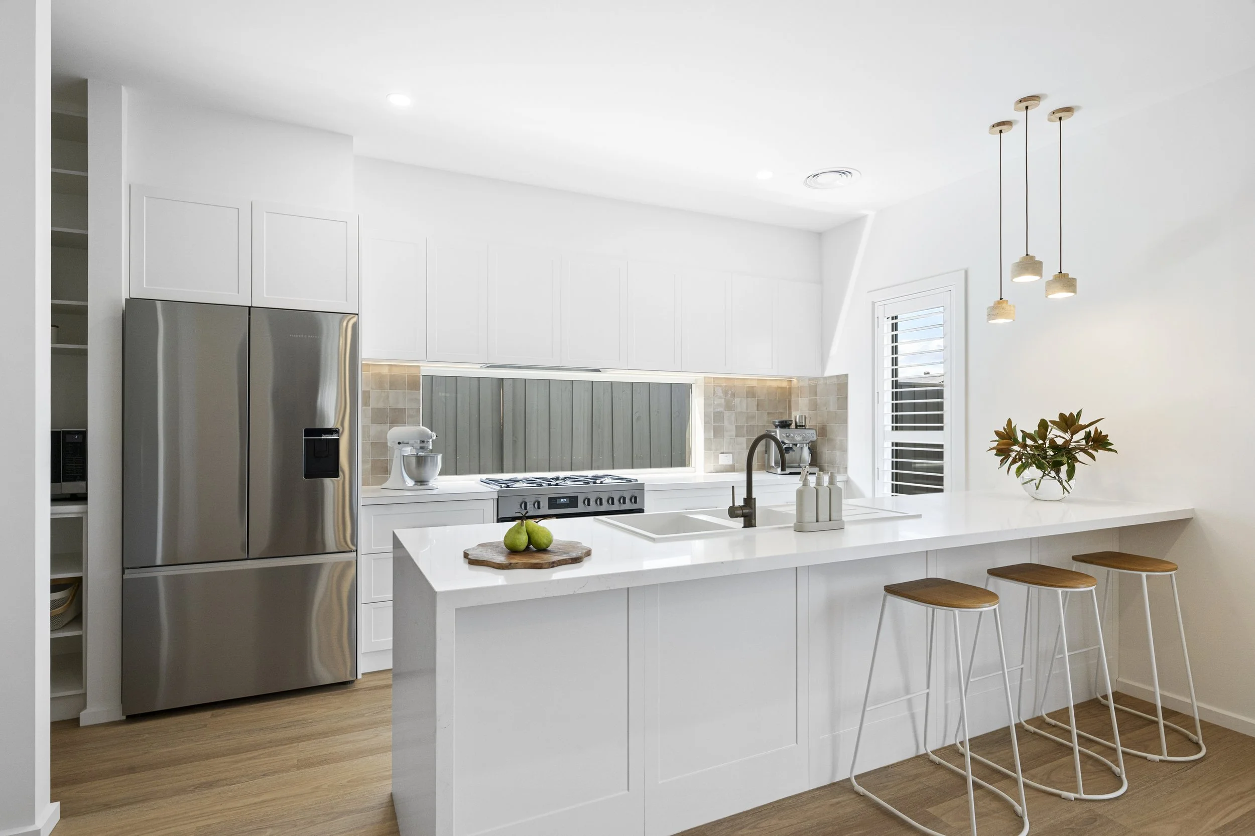 Modern white kitchen with a large island, three wooden bar stools, stainless steel refrigerator, and minimalistic decor.