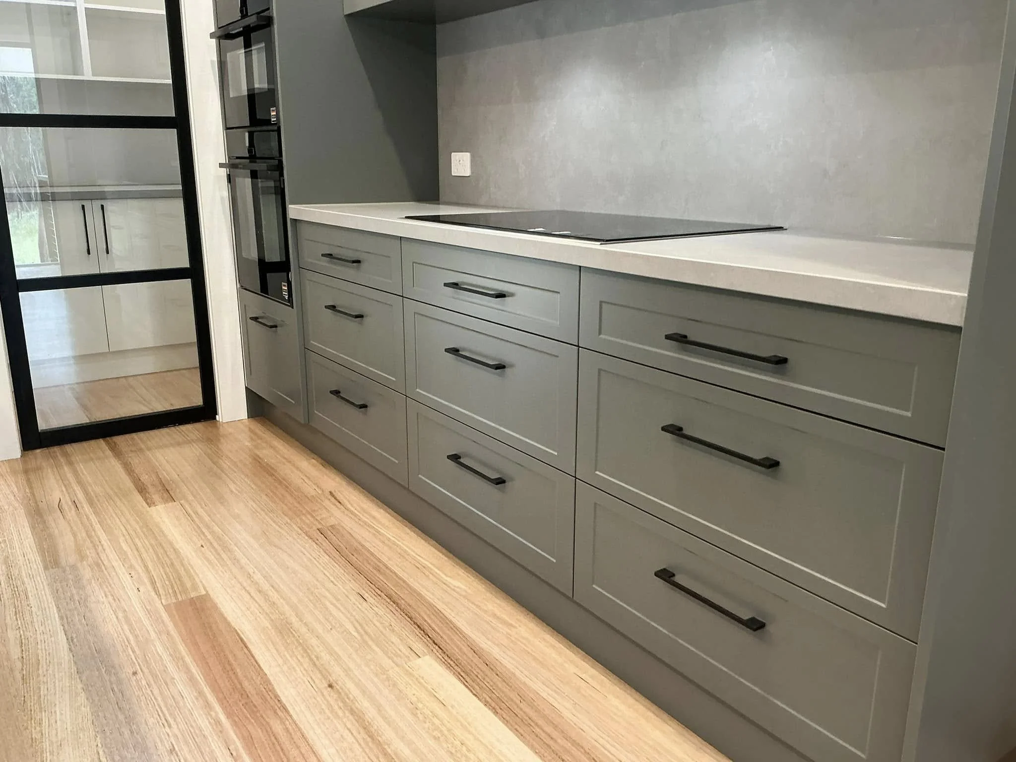 Modern kitchen with grey cabinets, black handles, white countertop, and wooden flooring. There is a built-in oven and a sleek stovetop.