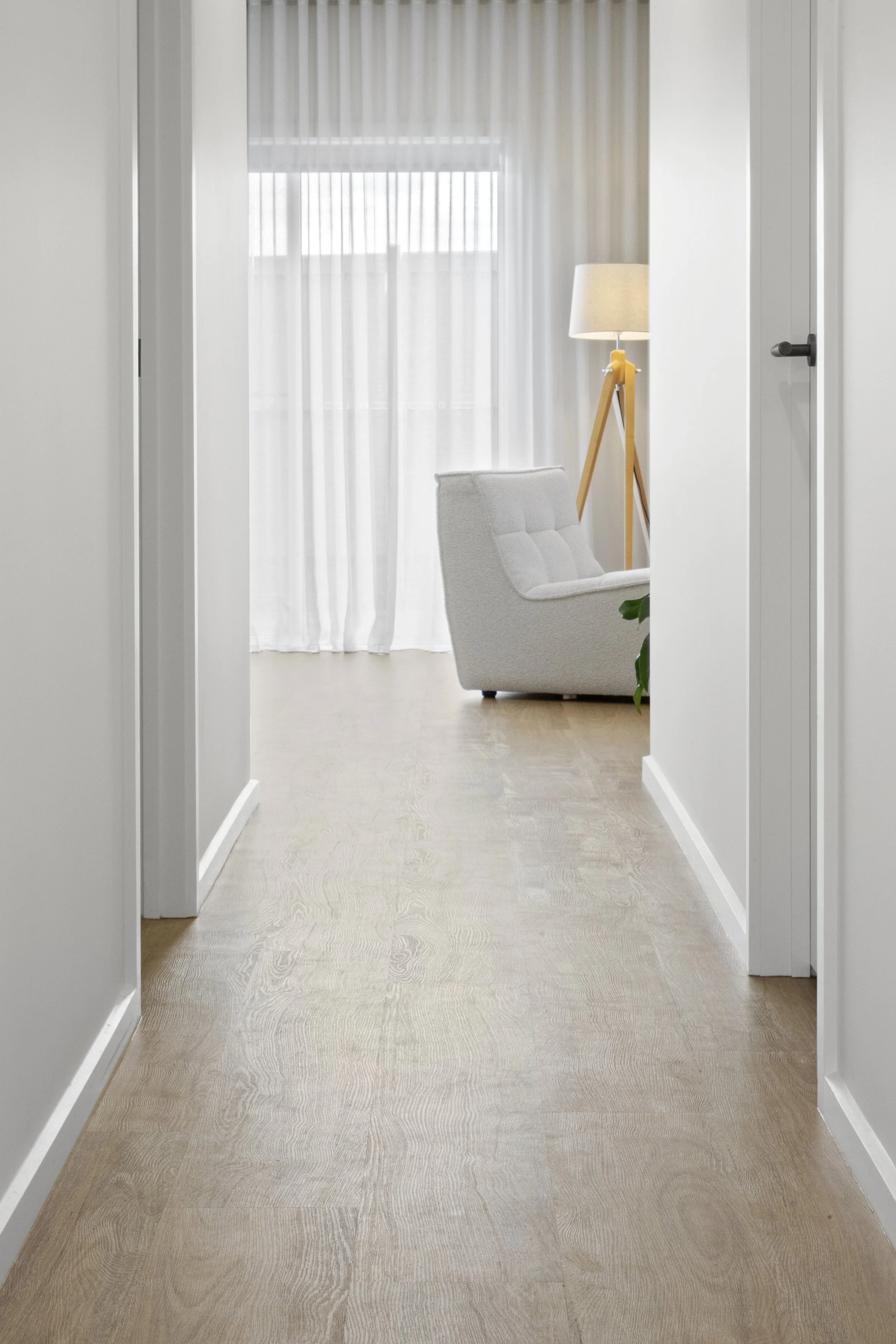 View through a hallway into a bright, white living room with sheer curtains, a white armchair, a floor lamp with a wooden tripod base, and a partial view of a plant.