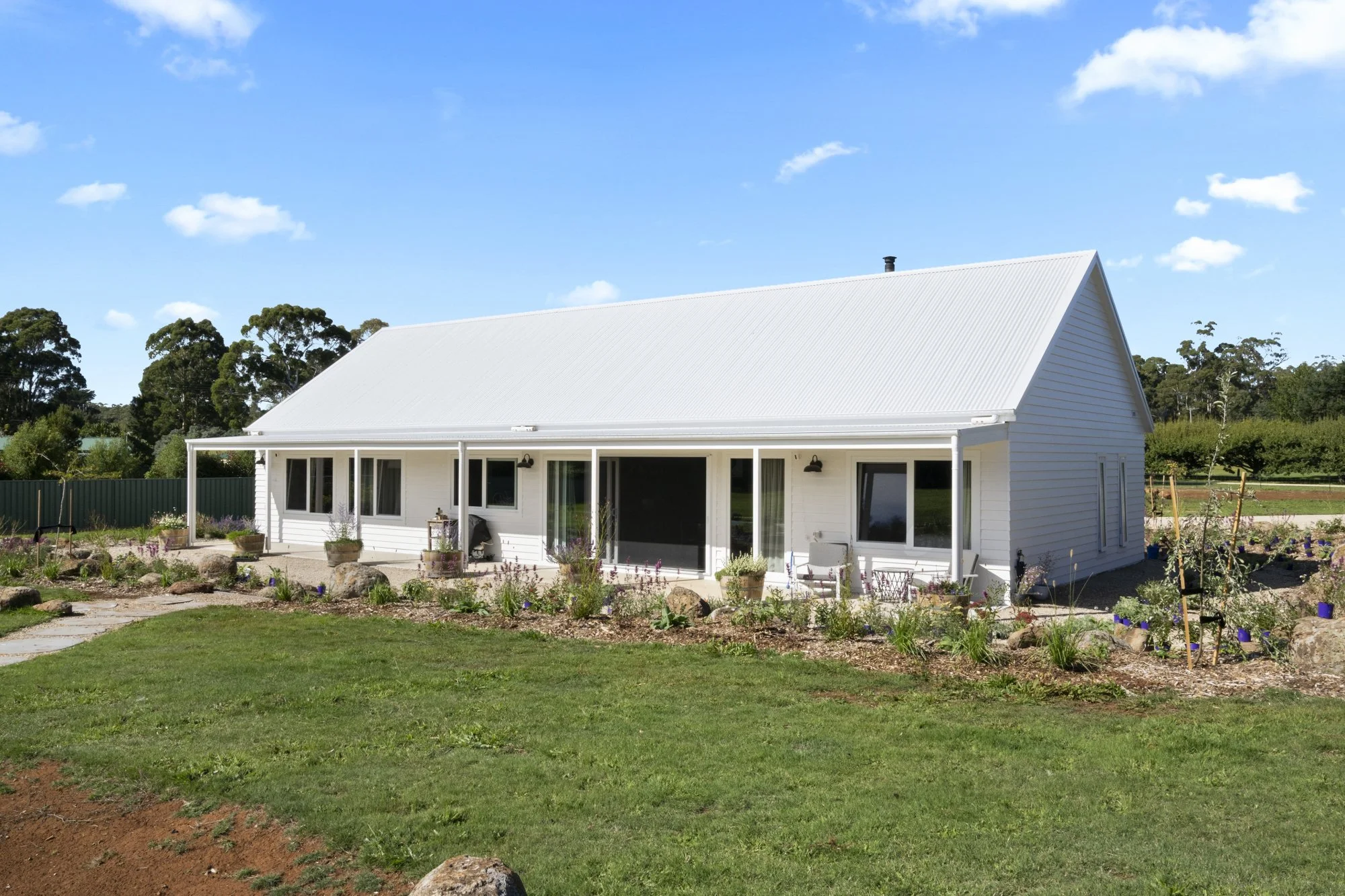 White house with a metal roof and a front porch, surrounded by a garden and grass lawn under a blue sky.