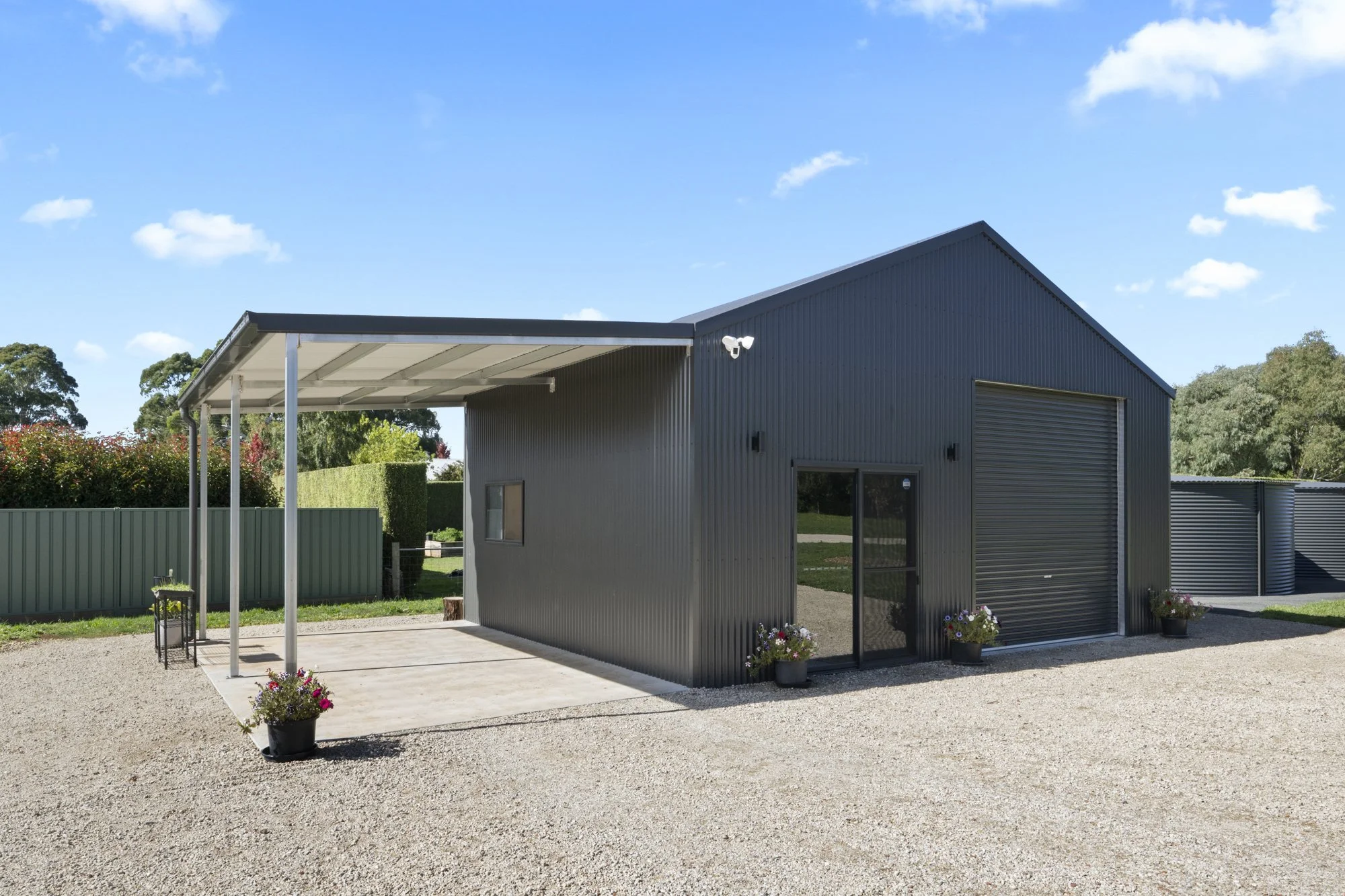 A modern metal building with a large roll-up door, a side entrance door with a screen door, and potted flowers outside. There is a gravel driveway and a small covered area supported by metal poles.