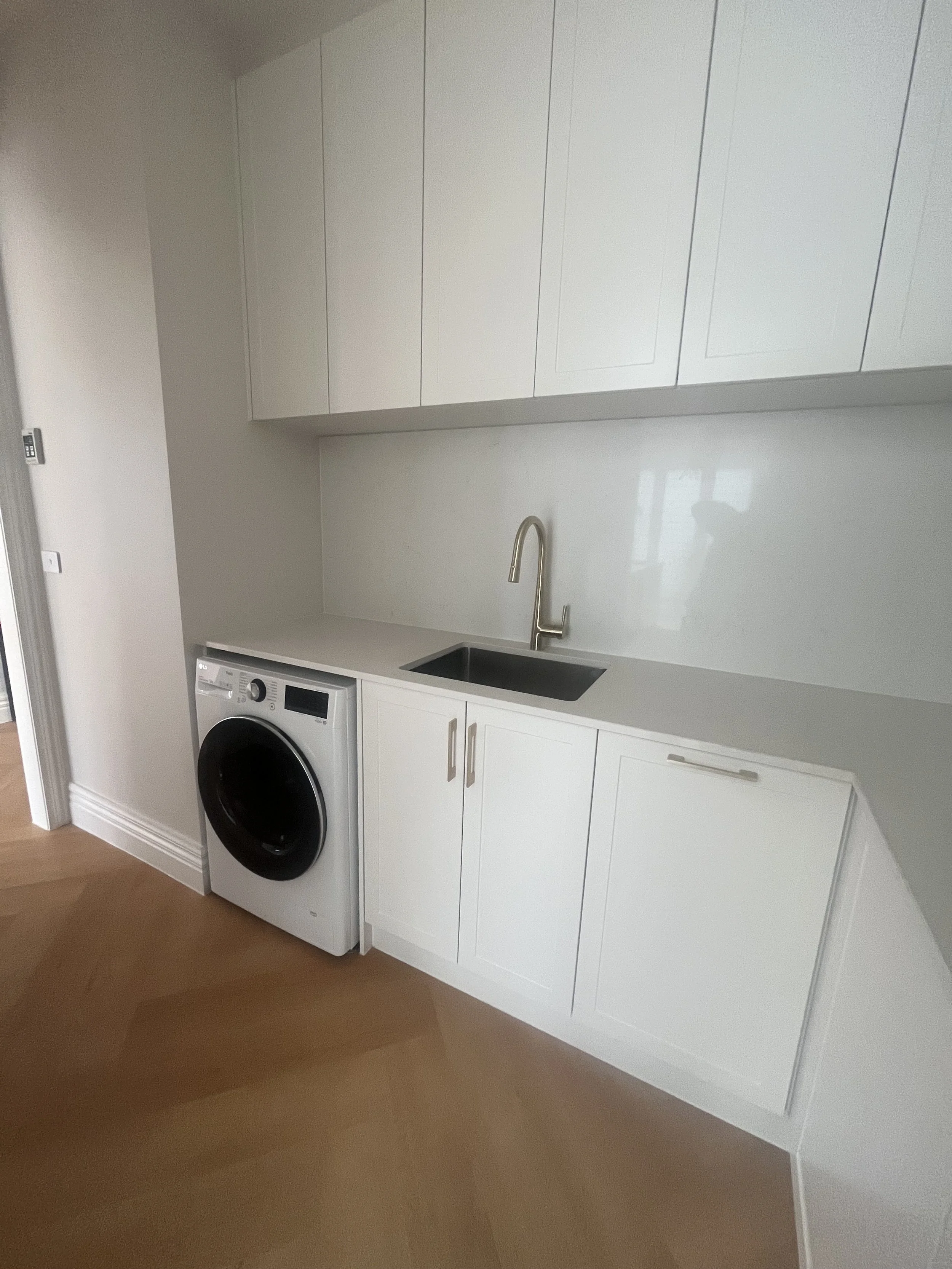 Modern laundry room with white cabinetry, a black front-loading washing machine, a stainless steel sink with a gold faucet, and light wooden flooring.