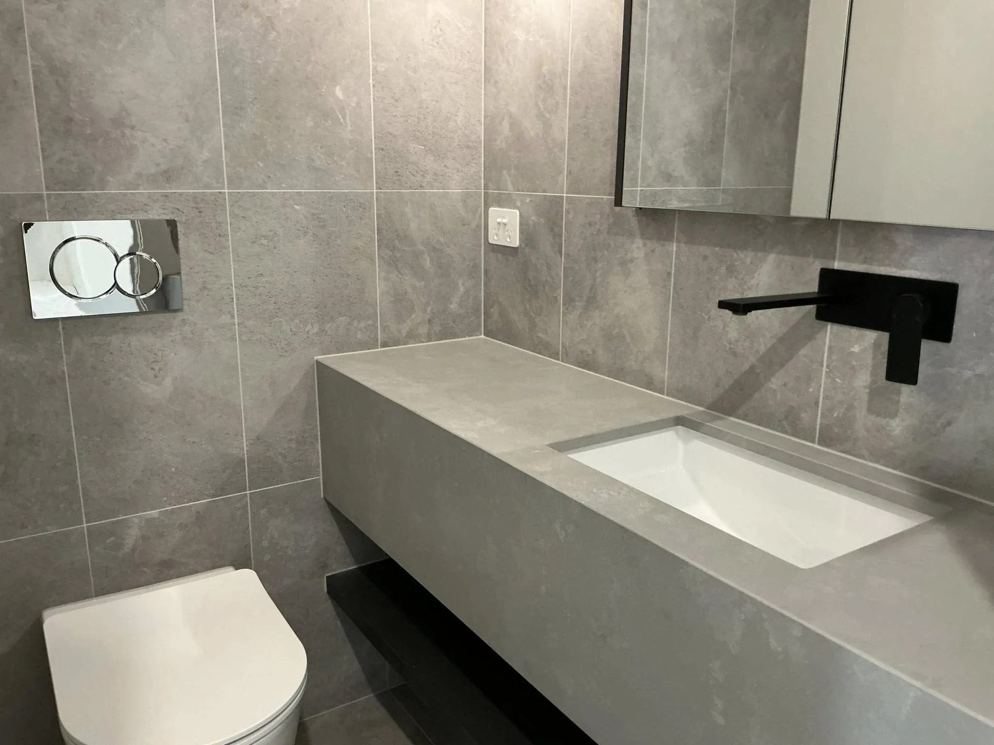 Modern bathroom with gray tiled walls and floor, a wall-mounted toilet with a concealed tank, a rectangular vanity with an integrated sink, a black faucet, a mirror cabinet, and a dual-ring flush plate on the wall.