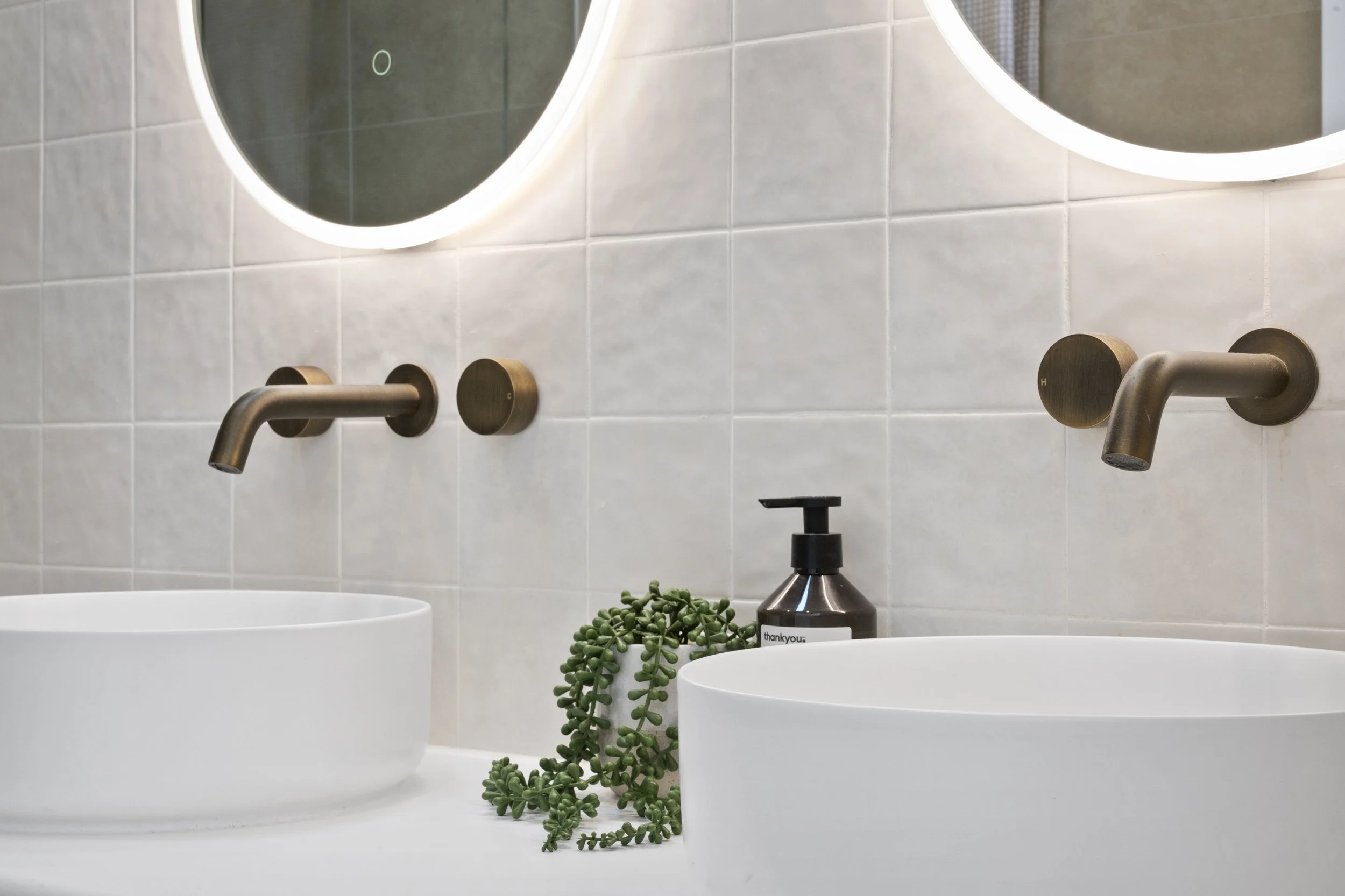 Modern bathroom sink with round white vessel basins, tan wall-mounted faucets, a black soap dispenser, and a decorative green plant, against a tiled wall with backlit mirrors.