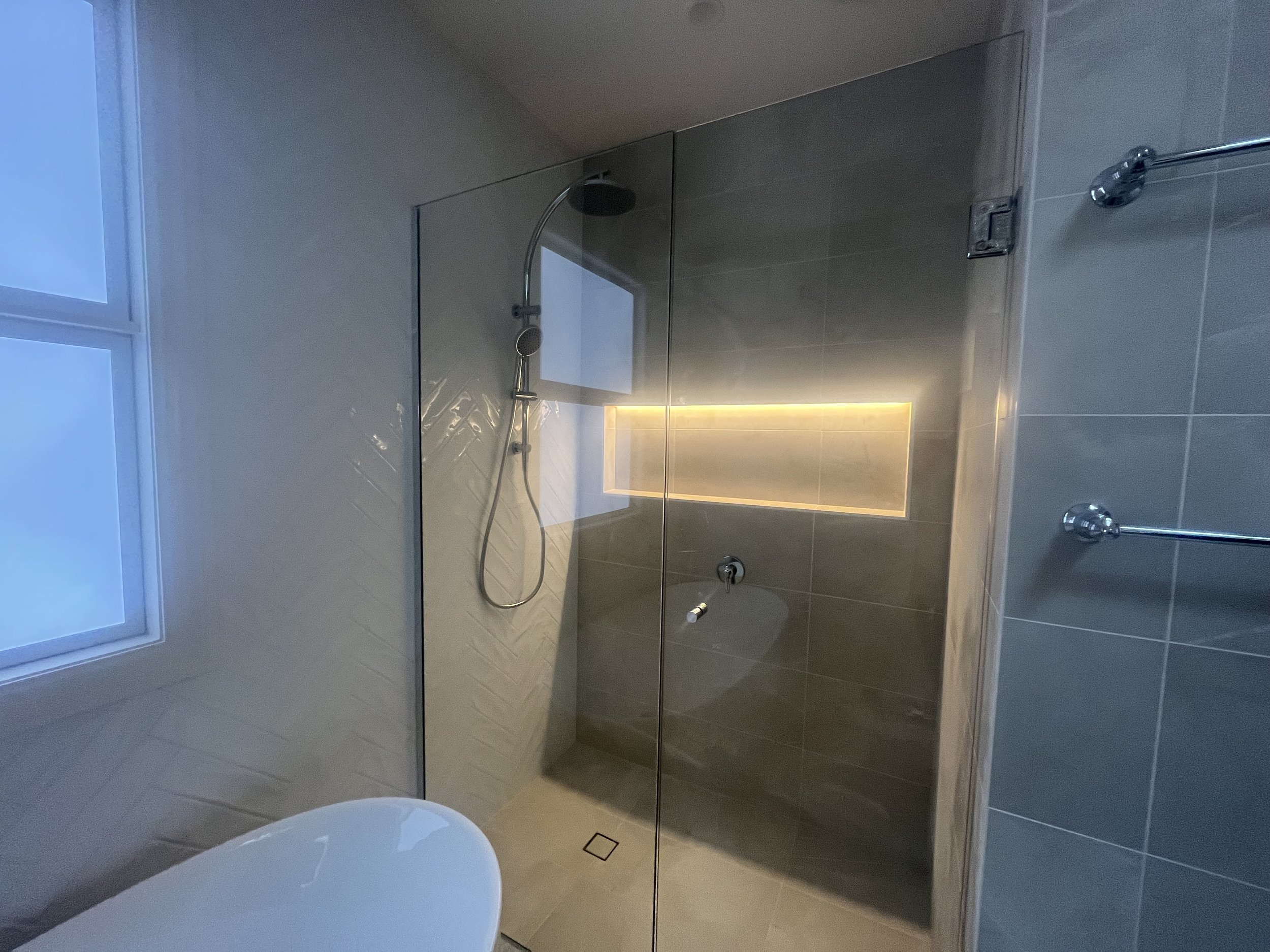 Modern bathroom shower area with glass enclosure, rainfall showerhead, and built-in wall niche with lighting, adjacent to a window.