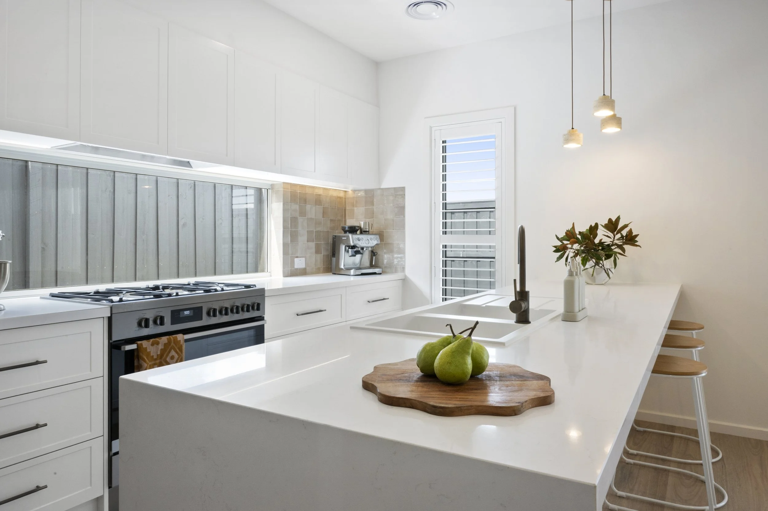 Modern kitchen with white cabinets, a white island with a built-in sink, a gas stove, a coffee machine, a vase with flowers, and three bar stools.