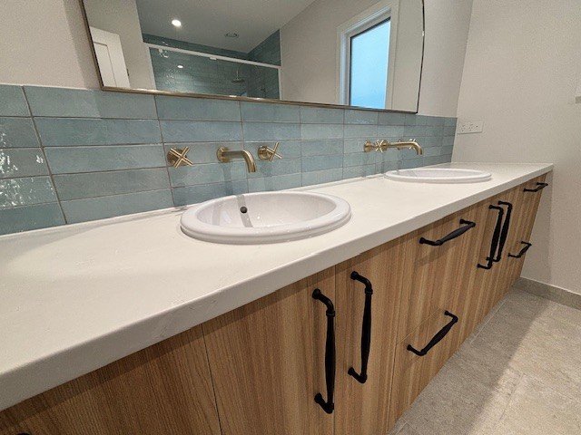 Bathroom with a double vanity, two sinks, brass faucets, a large mirror, light blue tile backsplash, and wood cabinets.