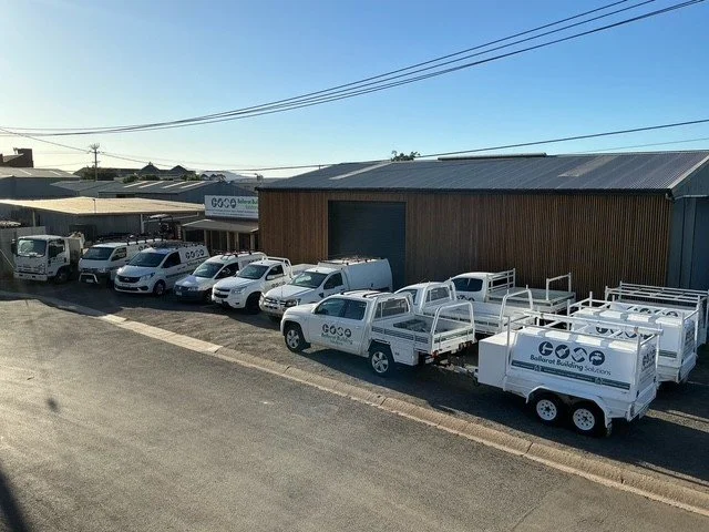 A row of white company vehicles, including utility trucks with flatbeds and small vans, parked in front of a wooden building during the daytime.