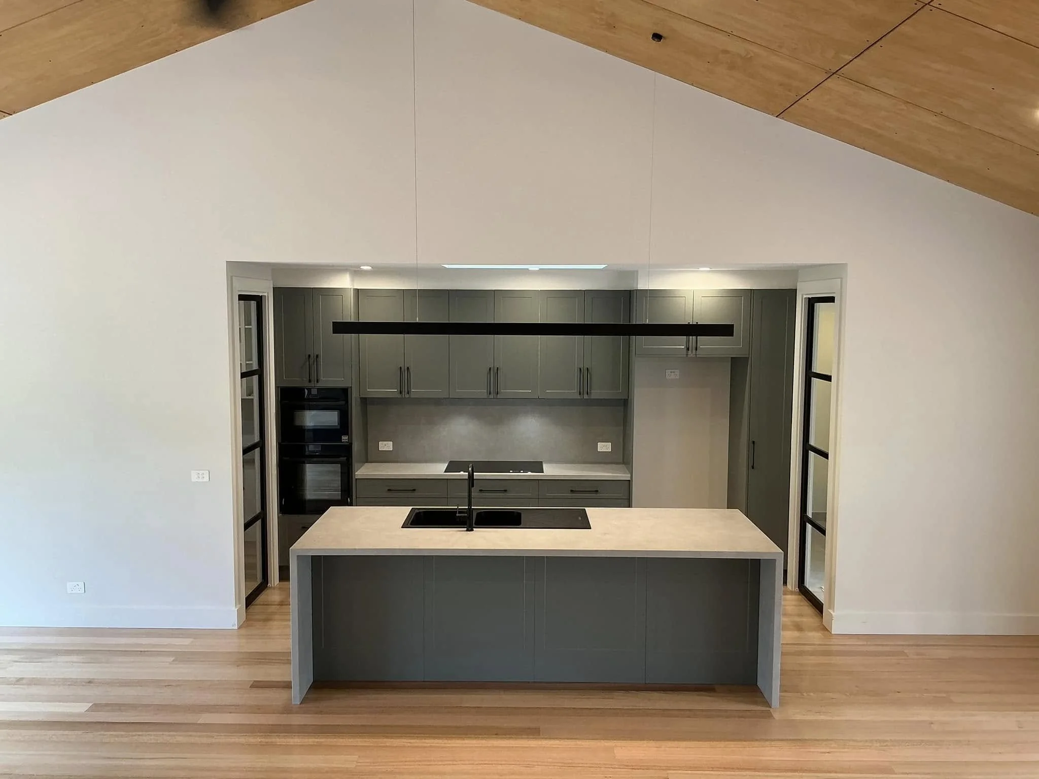 Modern kitchen with gray cabinets, a central island with a sink, and built-in appliances, viewed from a room with white walls and wooden floors.