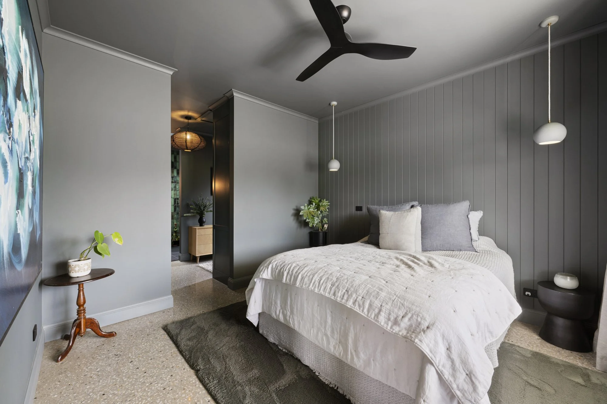 Modern bedroom with a bed featuring various pillows, dark gray accent wall behind it, pendant lights hanging on either side, black ceiling fan, and small side tables with plants.