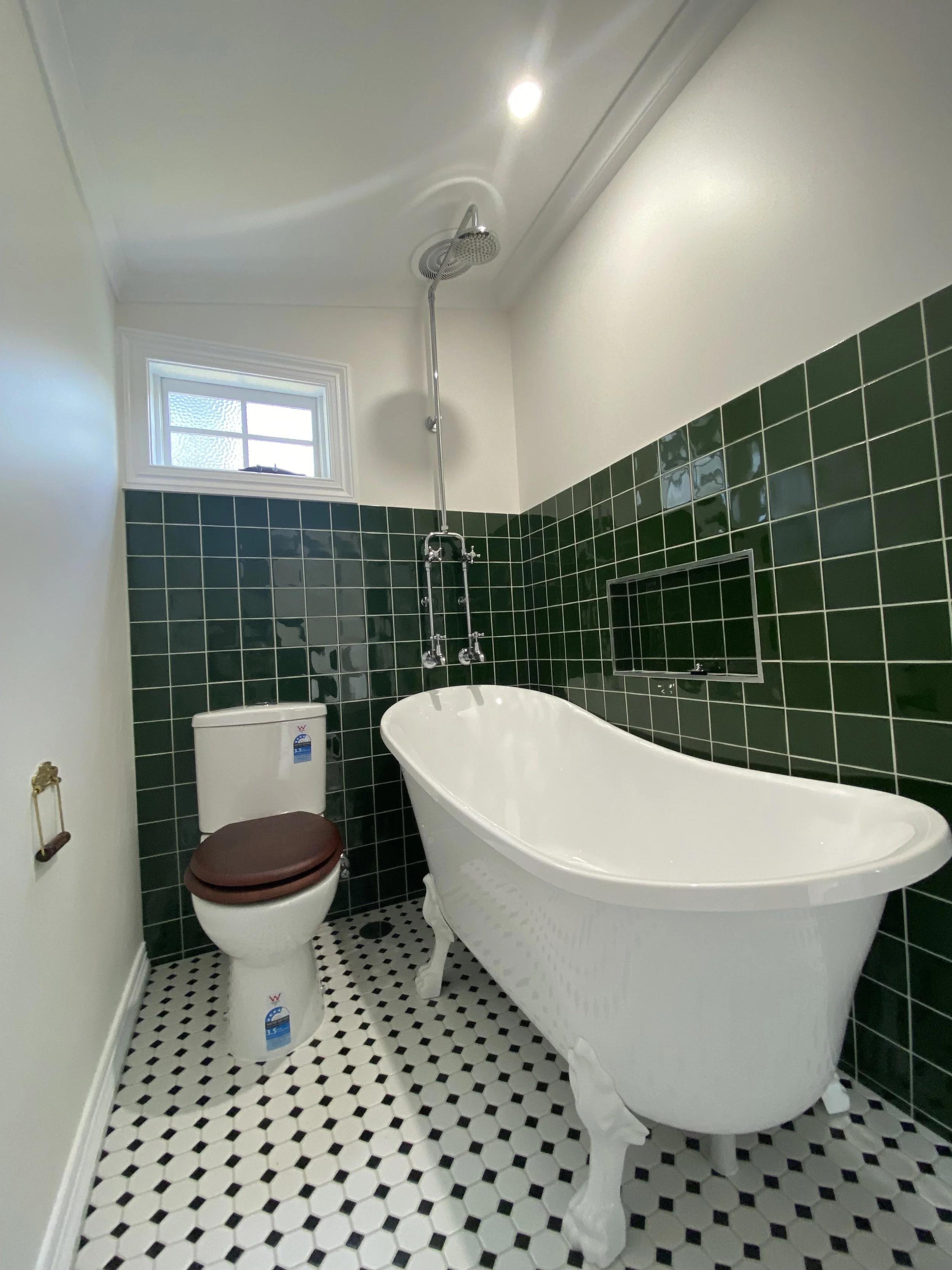 A bathroom with green tiled walls, a white clawfoot bathtub, a brown toilet seat, a small window, and black and white hexagonal floor tiles.