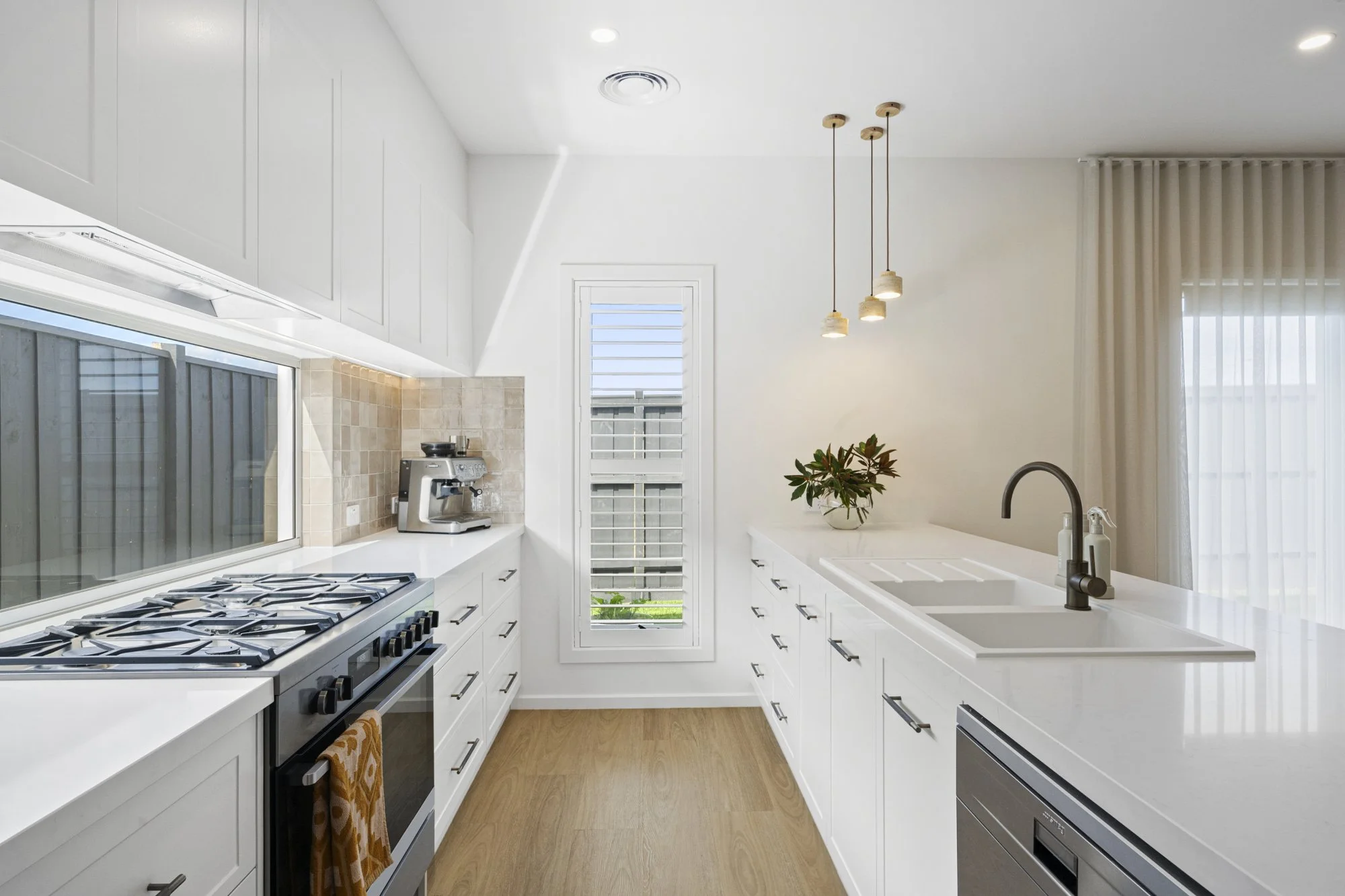 Modern white kitchen with gas stove, built-in microwave, coffee machine, white countertops, upper and lower cabinets, a window, pendant lights, a potted plant, and a sink with a black faucet.