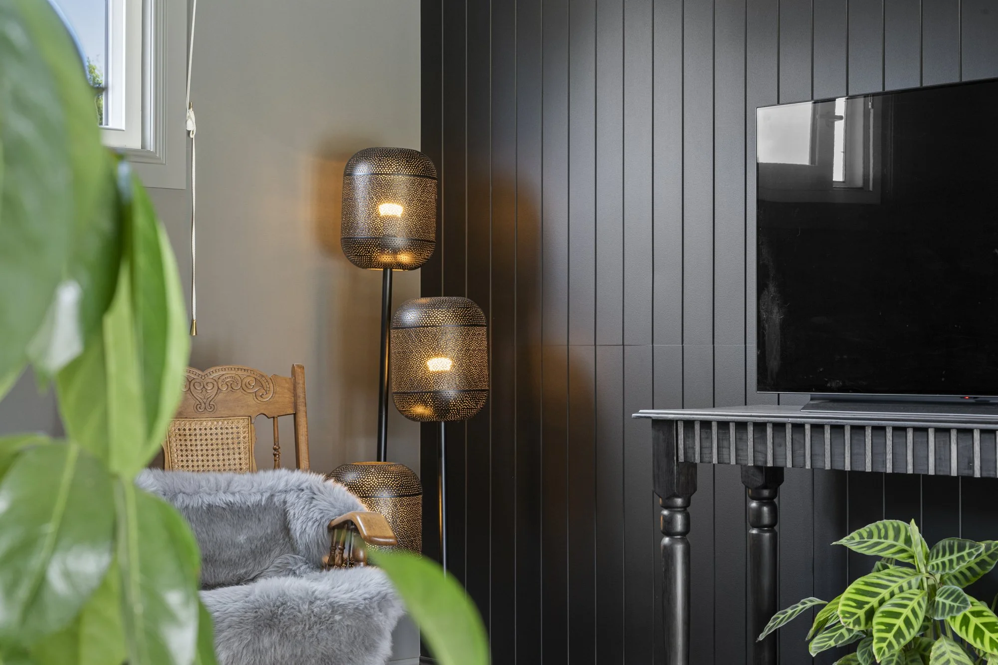 Living room corner with a wooden chair with gray faux fur throw, black wall with vertical panels, a modern black floor lamp with three perforated metal shades, a TV on a black table, and green houseplants near the window.