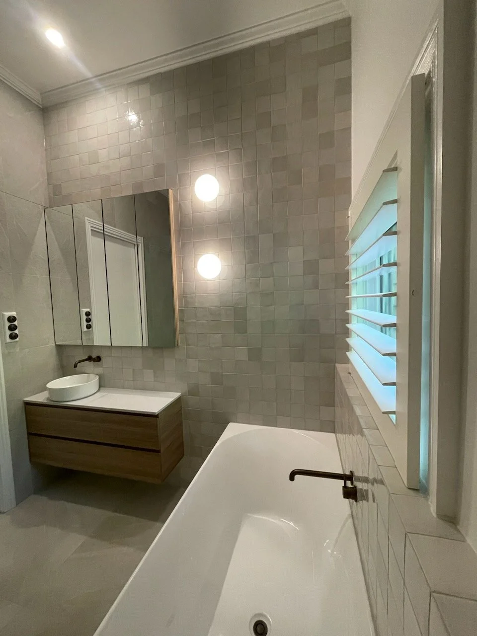 Modern bathroom with bathtub, wall-mounted vanity with vessel sink, mirror cabinet, window with shutters, and beige tiled walls.