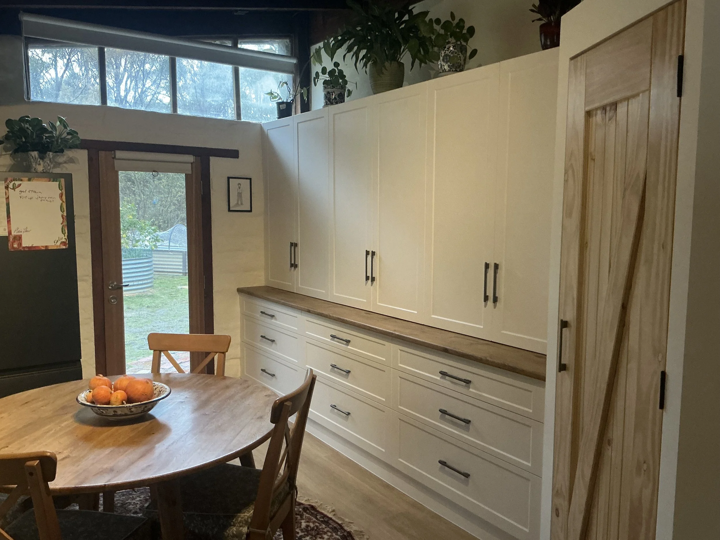 A kitchen corner with a white cabinet, wooden countertop, and a wooden sliding door. A wooden dining table with a bowl of fruit and a wooden chair is in the foreground. Large windows and a door lead to a backyard.
