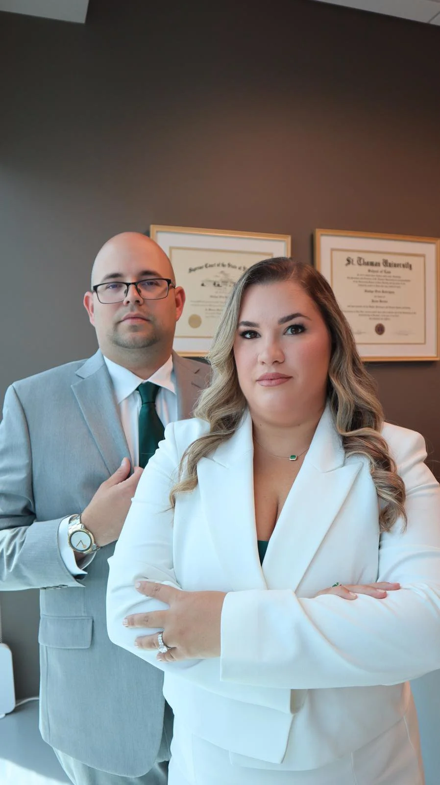 A male attorney and female attorney, dressed professionally, standing together in an office with diplomas on the wall behind them.