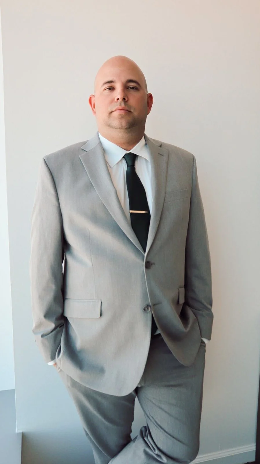 A male attorney in a light gray suit, white shirt, black tie, standing with one hand in his pocket against a plain wall.