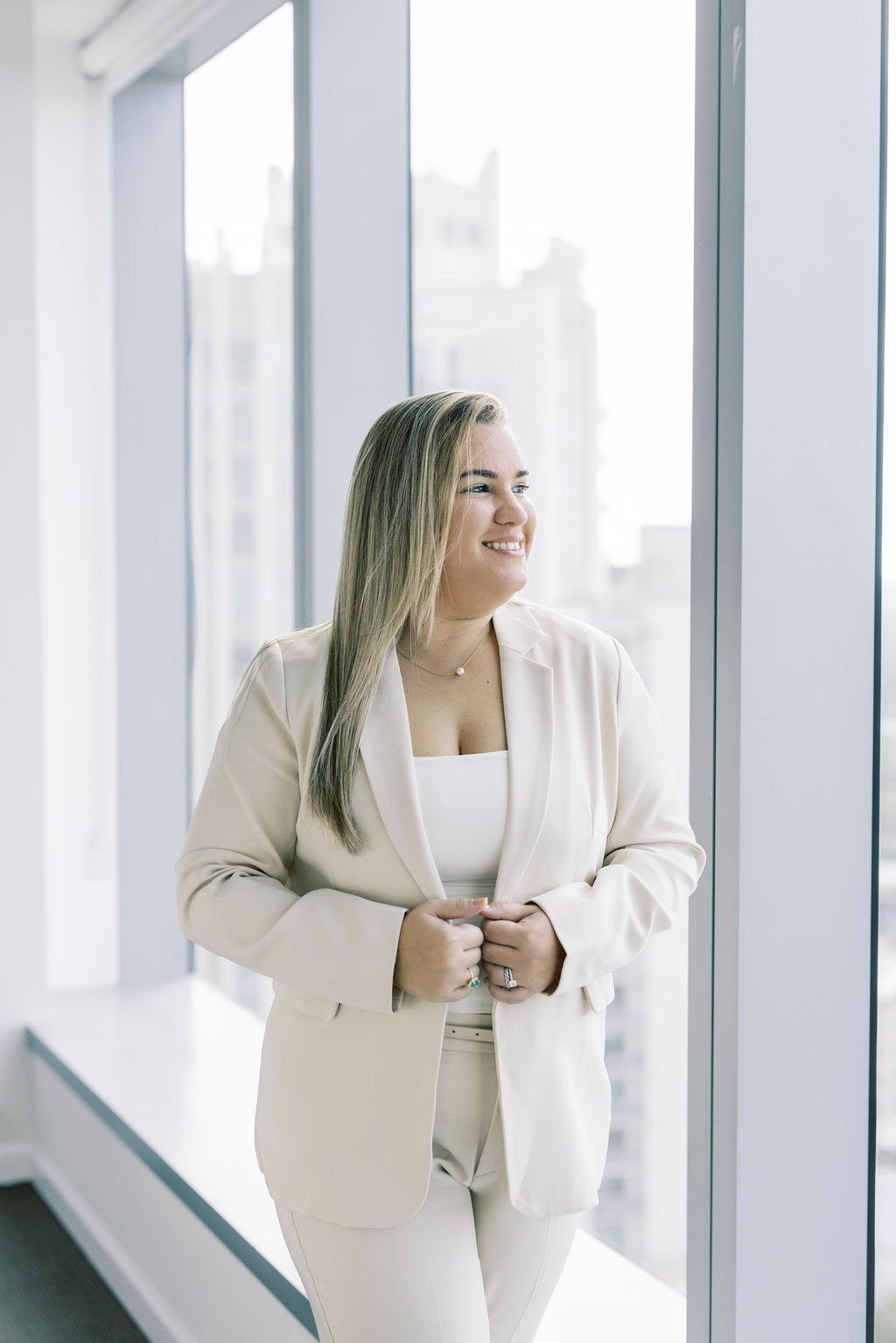 A woman with long blonde hair smiling and looking out a large window in a modern office building. She is wearing a white blazer and matching pants.